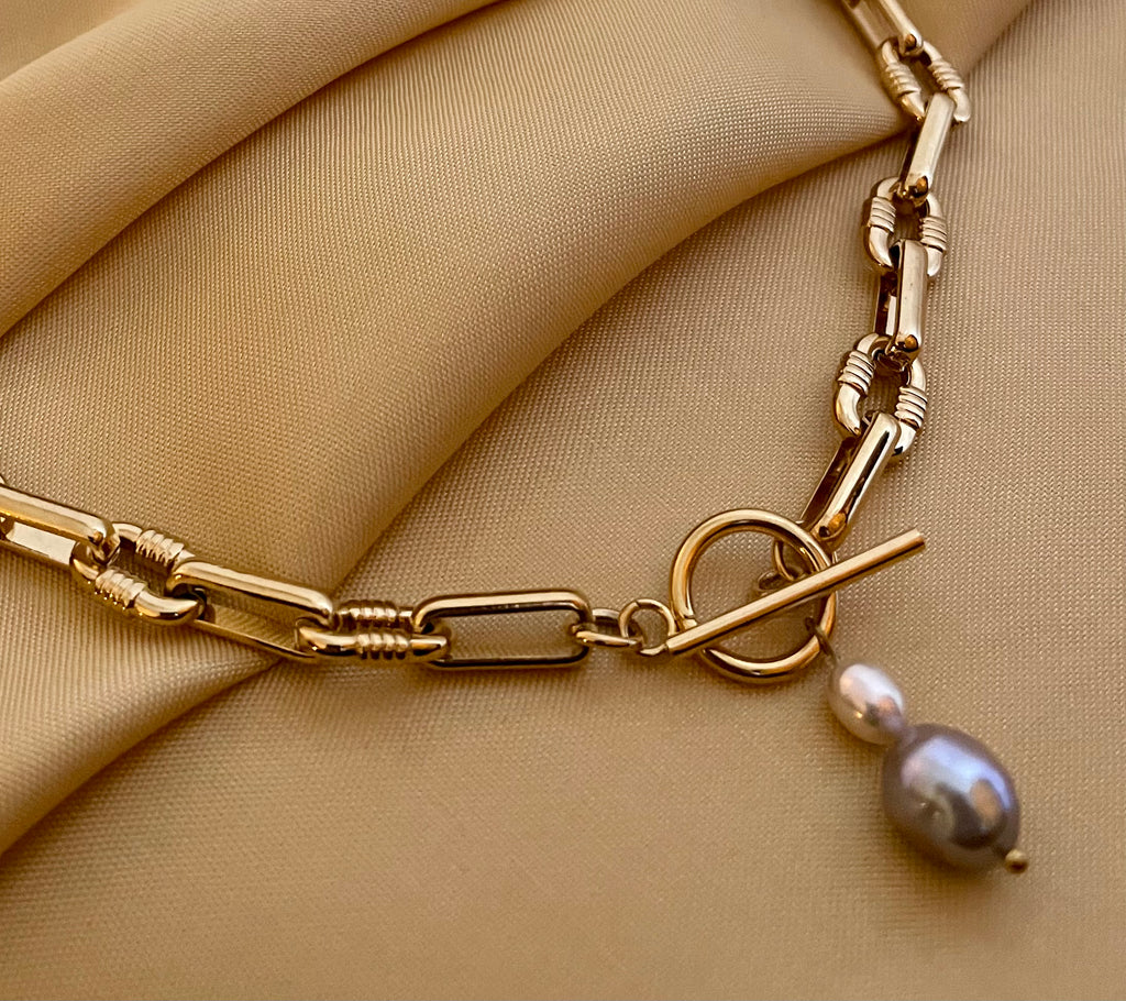 Detail shot of a luxury gold link necklace worn on an ivory silk top. The focal point is a T-bar toggle closure holding a high-shine metallic grey baroque pearl and a champagne oval pearl, showing deep luster and smooth nacre
