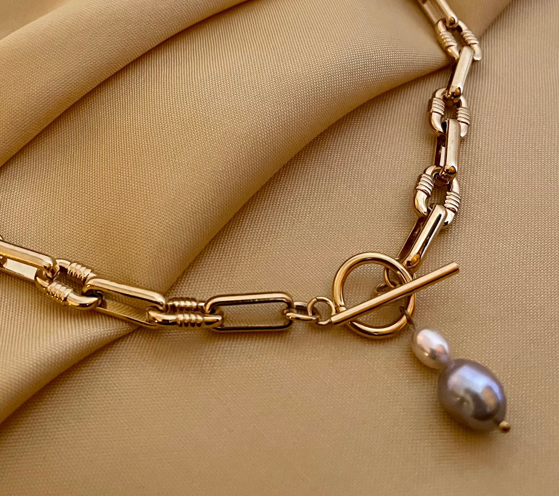 Detail shot of a luxury gold link necklace worn on an ivory silk top. The focal point is a T-bar toggle closure holding a high-shine metallic grey baroque pearl and a champagne oval pearl, showing deep luster and smooth nacre