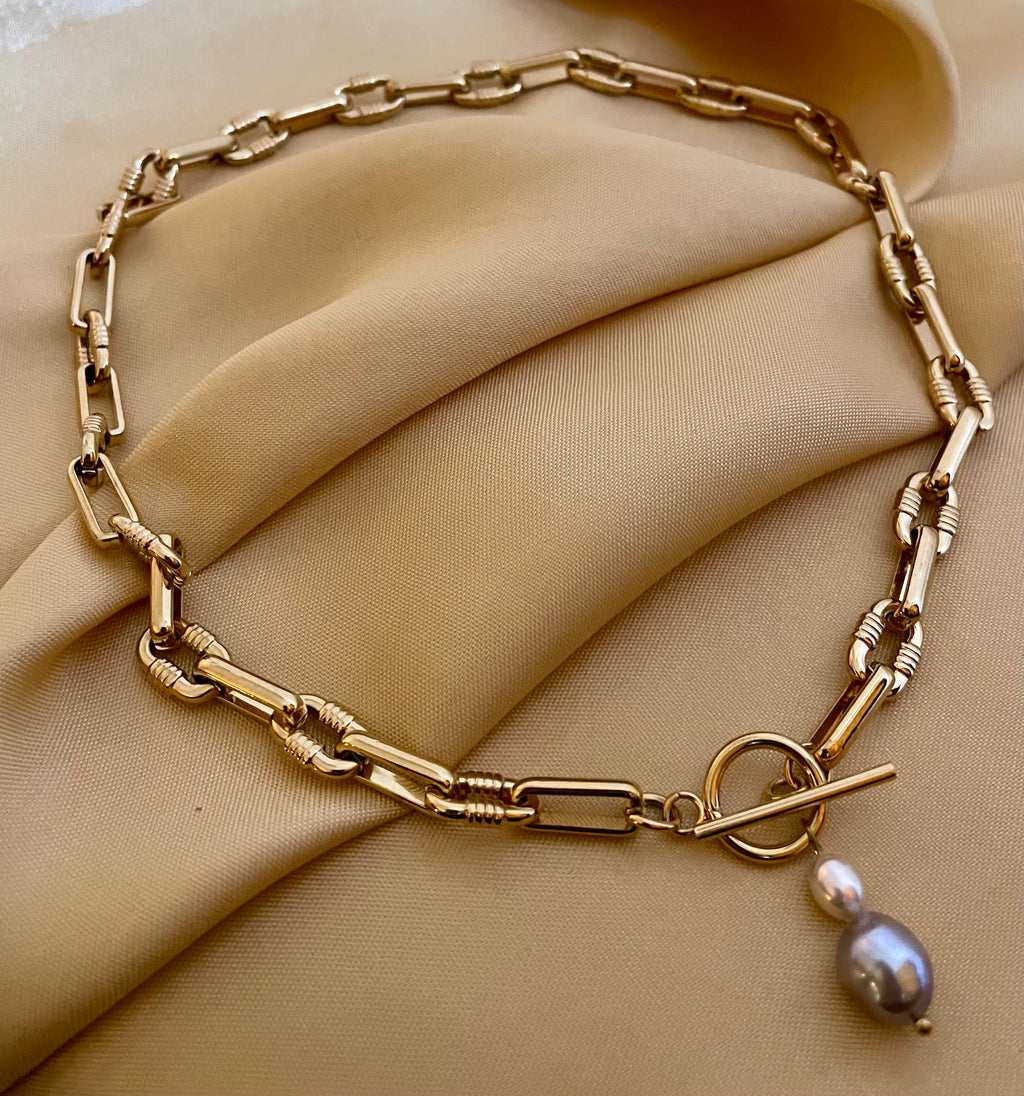 Detail shot of a luxury gold link necklace. The focal point is a T-bar toggle closure holding a high-shine metallic grey baroque pearl and a champagne oval pearl, showing deep luster and smooth nacre