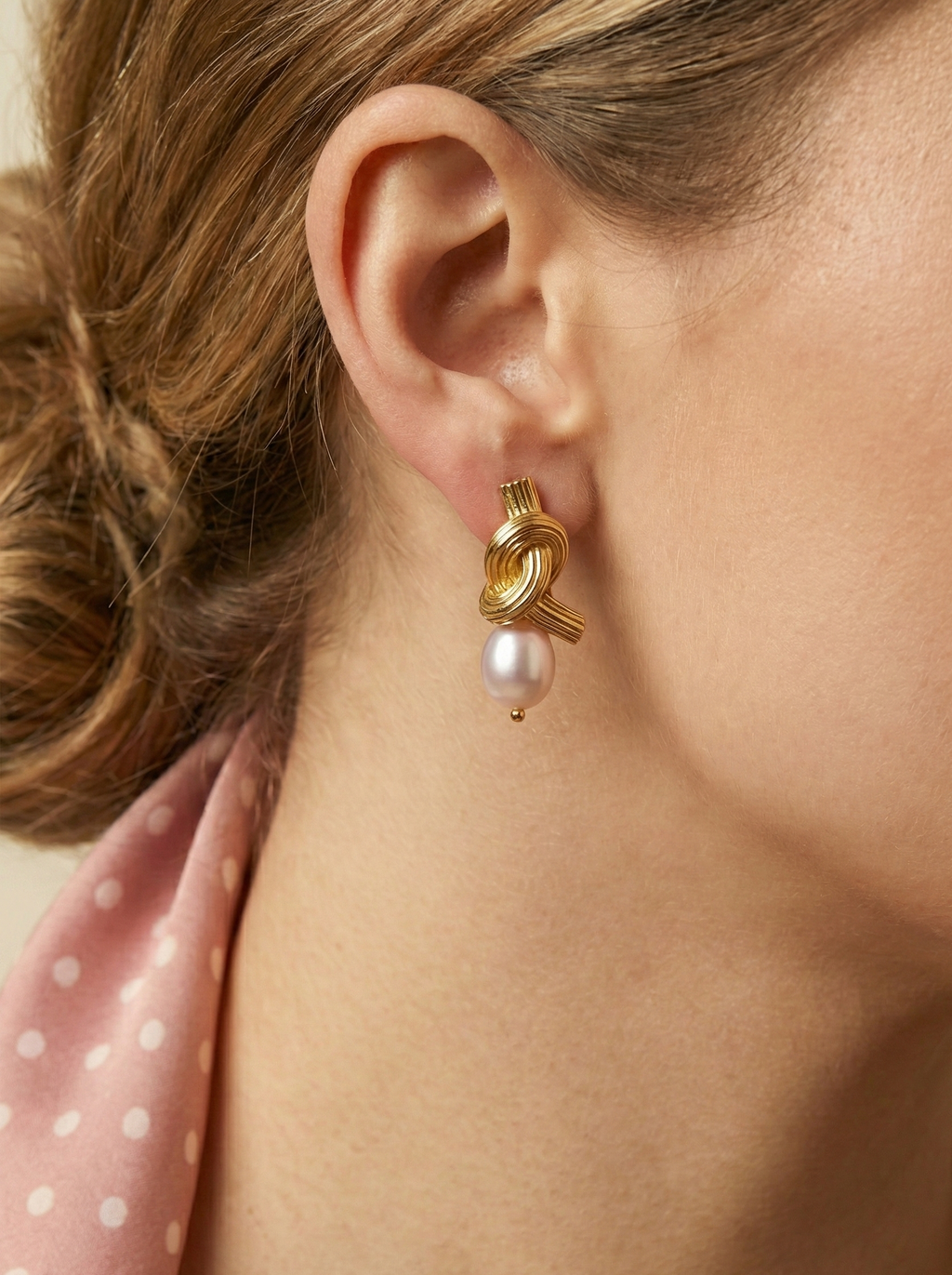 Close-up editorial photography of ribbed gold twisted knot earrings featuring a lustrous pink oval pearl drop. Modeled on a woman with a low bun hairstyle wearing a silk olive camisole and pink neck scarf, highlighting the earring's luxe texture and elegant drape.