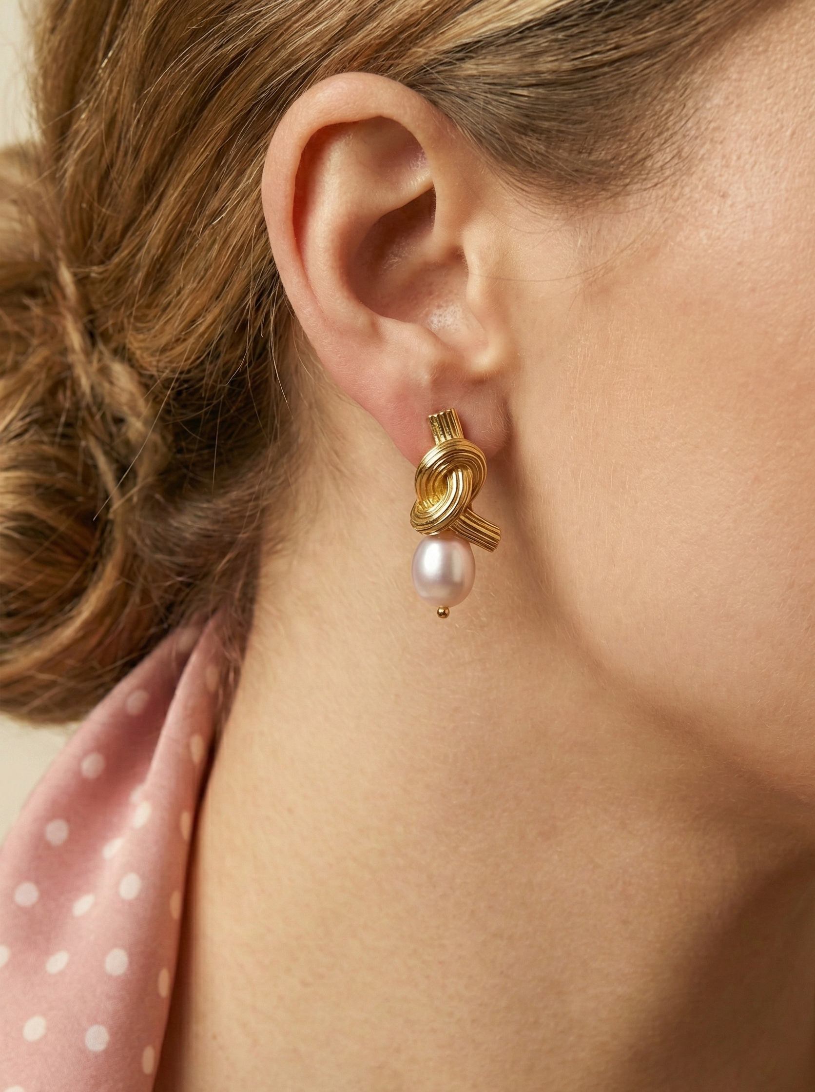 Close-up editorial photography of ribbed gold twisted knot earrings featuring a lustrous pink oval pearl drop. Modeled on a woman with a low bun hairstyle wearing a silk olive camisole and pink neck scarf, highlighting the earring's luxe texture and elegant drape.