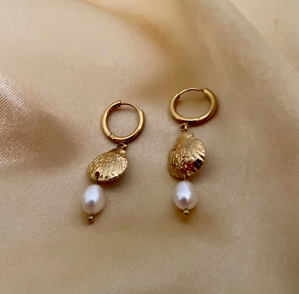 French style inspiration: A model with blonde-brown hair in a messy low bun wearing textured gold sea shell huggie earrings with a dainty pearl charm. Soft studio lighting highlights the iridescence of the pearl and the model's mauve camisole