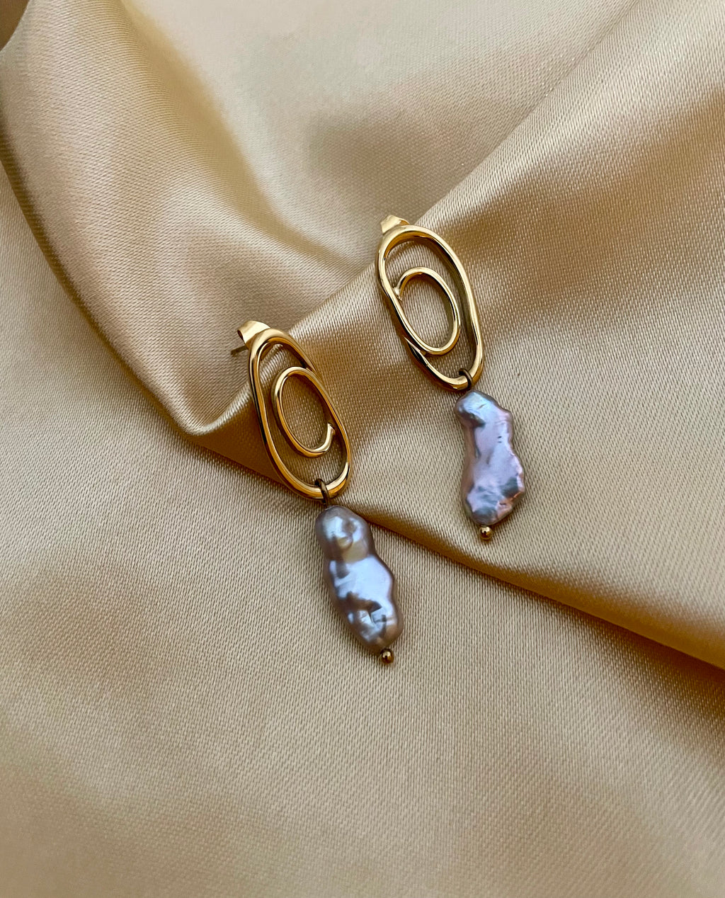 Close-up of a  luxury grey baroque keshi pearl drop earrings with a sculptural gold double-oval link stud. The image highlights the iridescent nacre and organic shape of the pearl, styled against a soft matte ivory background, representing high-end commercial jewelry photography
