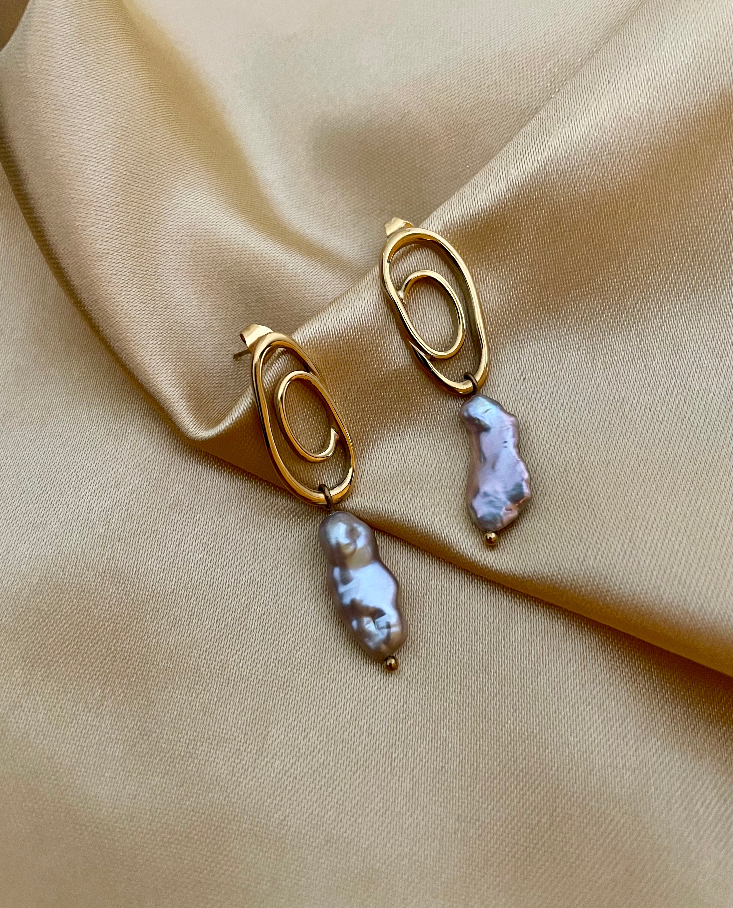 Close-up of a  luxury grey baroque keshi pearl drop earrings with a sculptural gold double-oval link stud. The image highlights the iridescent nacre and organic shape of the pearl, styled against a soft matte ivory background, representing high-end commercial jewelry photography