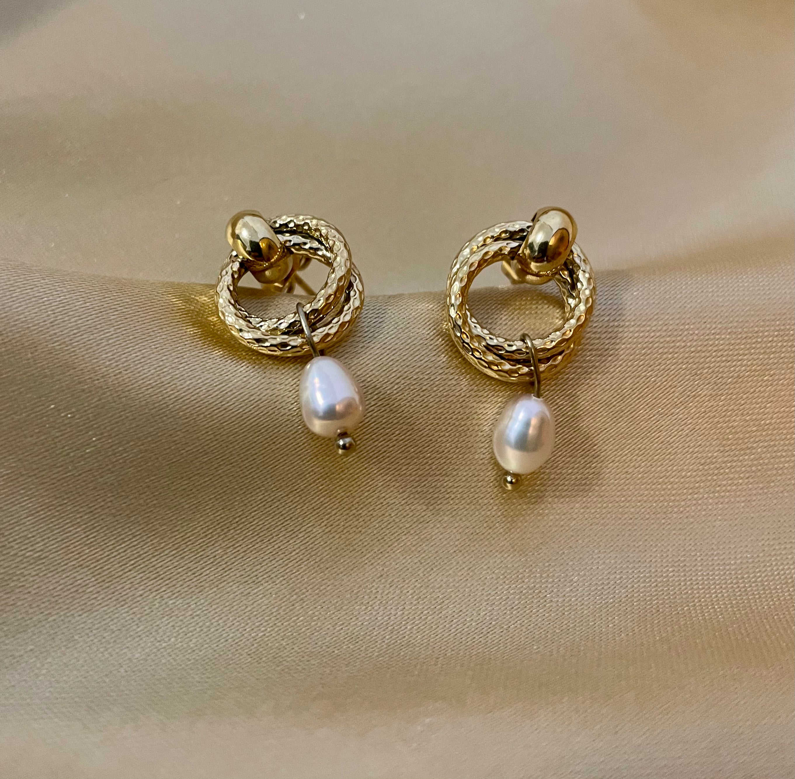 Close-up detail of textured gold hoop earrings featuring a dainty oval pearl drop. The earrings are worn on the ear to show scale, highlighting the metallic luster and elegant Parisian chic design against a neutral background.