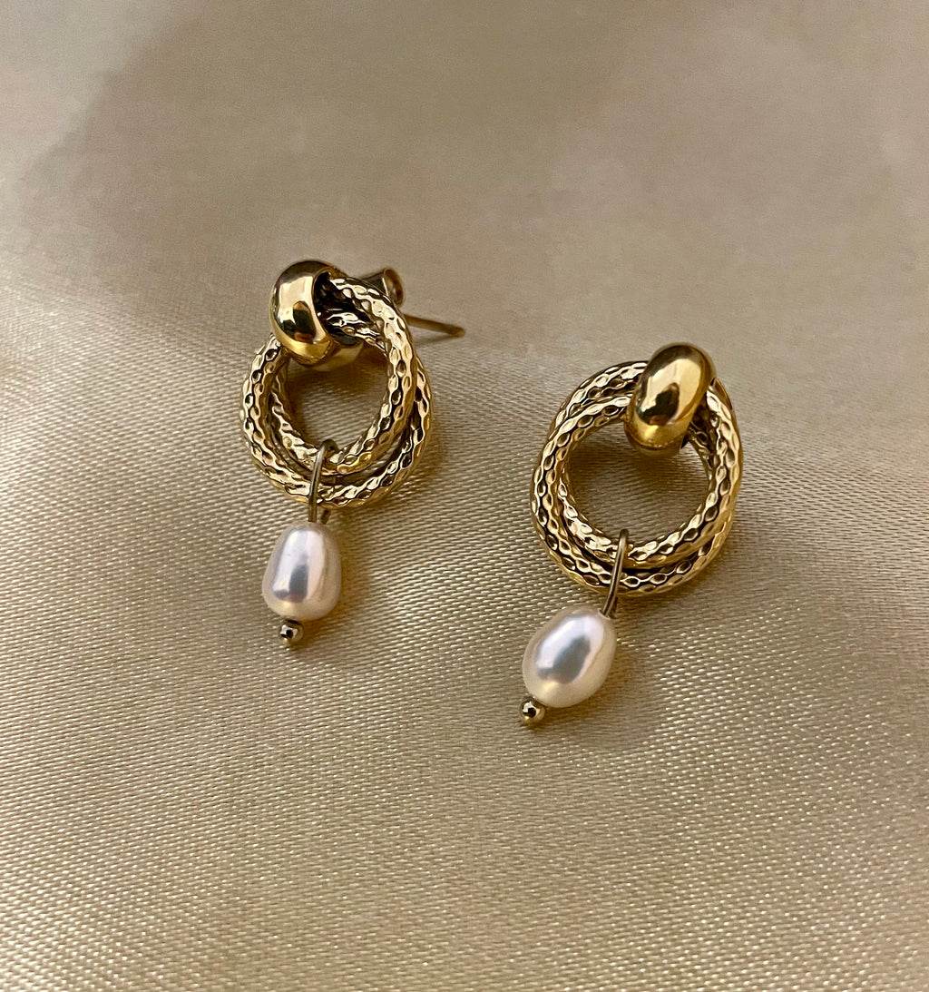 Close-up detail of textured gold hoop earrings featuring a dainty oval pearl drop. The earrings are worn on the ear to show scale, highlighting the metallic luster and elegant Parisian chic design against a neutral background.