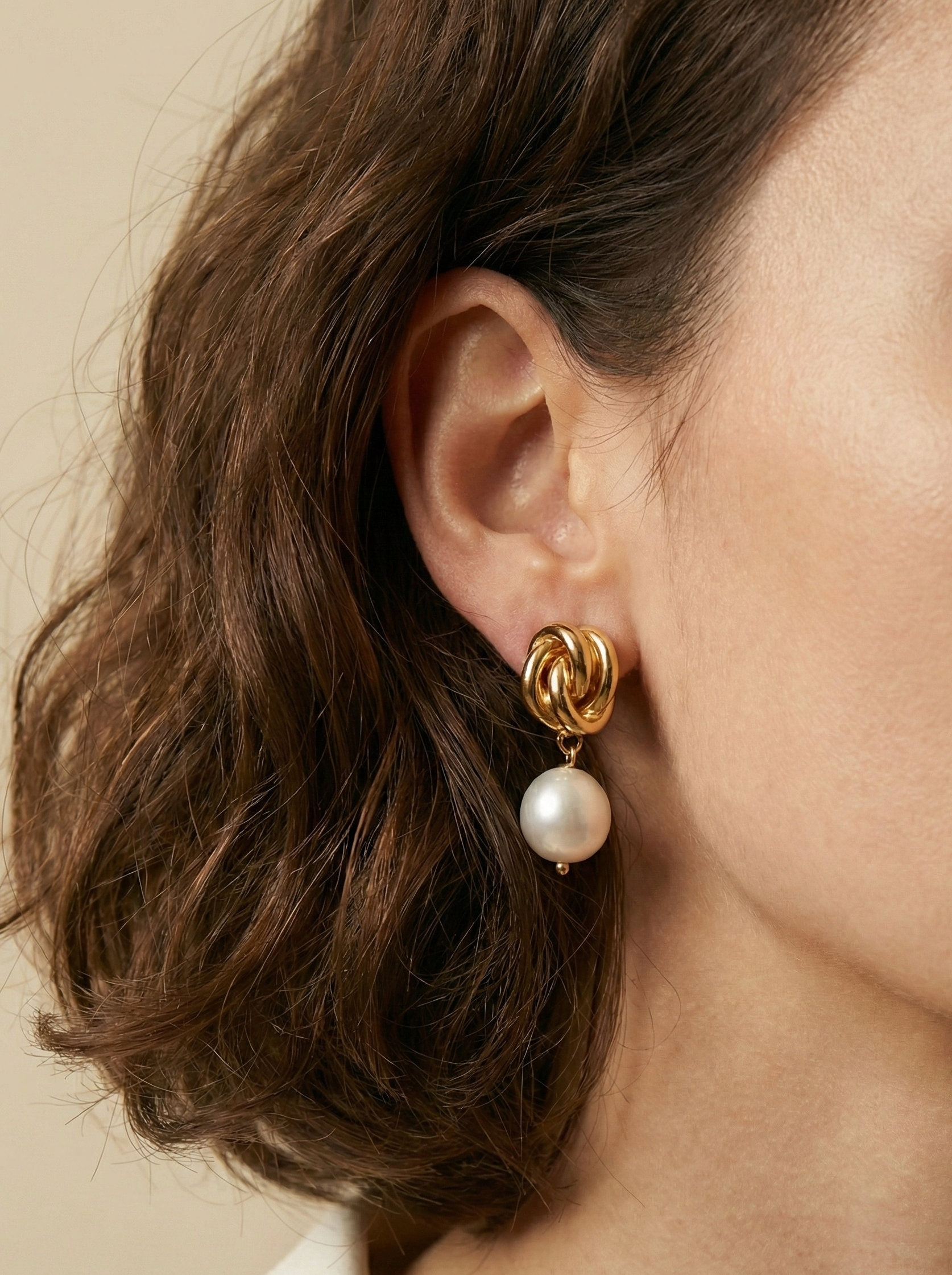Close-up detail of a woman's ear wearing gold twist-knot stud earrings with a lustrous white pearl drop. The lighting highlights the texture of the gold and the glow of the pearl