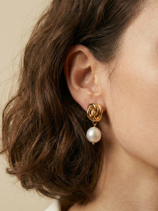 Close-up detail of a woman's ear wearing gold twist-knot stud earrings with a lustrous white pearl drop. The lighting highlights the texture of the gold and the glow of the pearl