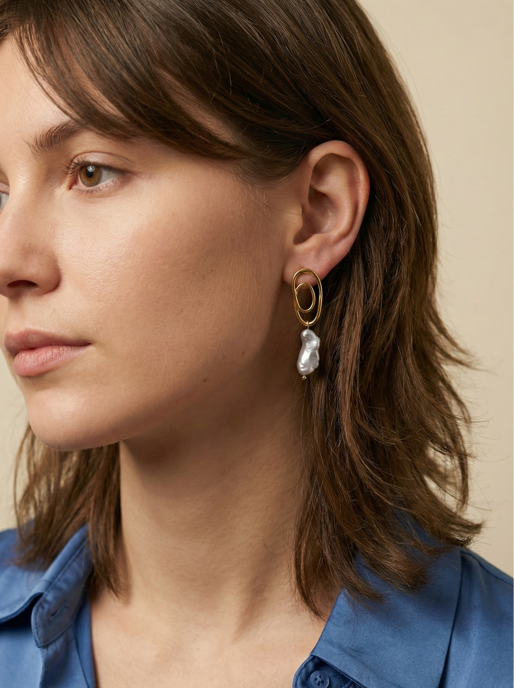 Close-up profile of a Korean model wearing luxury grey baroque pearl drop earrings with a sculptural gold double-oval link stud. The image highlights the iridescent nacre and organic shape of the pearl, styled with a blue knit top against a soft matte ivory background, representing high-end commercial jewelry photography