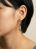 Close-up profile of a french model wearing luxury grey baroque pearl drop earrings with a sculptural gold double-oval link stud. The image highlights the iridescent nacre and organic shape of the pearl, styled with a blue knit top against a soft matte ivory background, representing high-end commercial jewelry photography