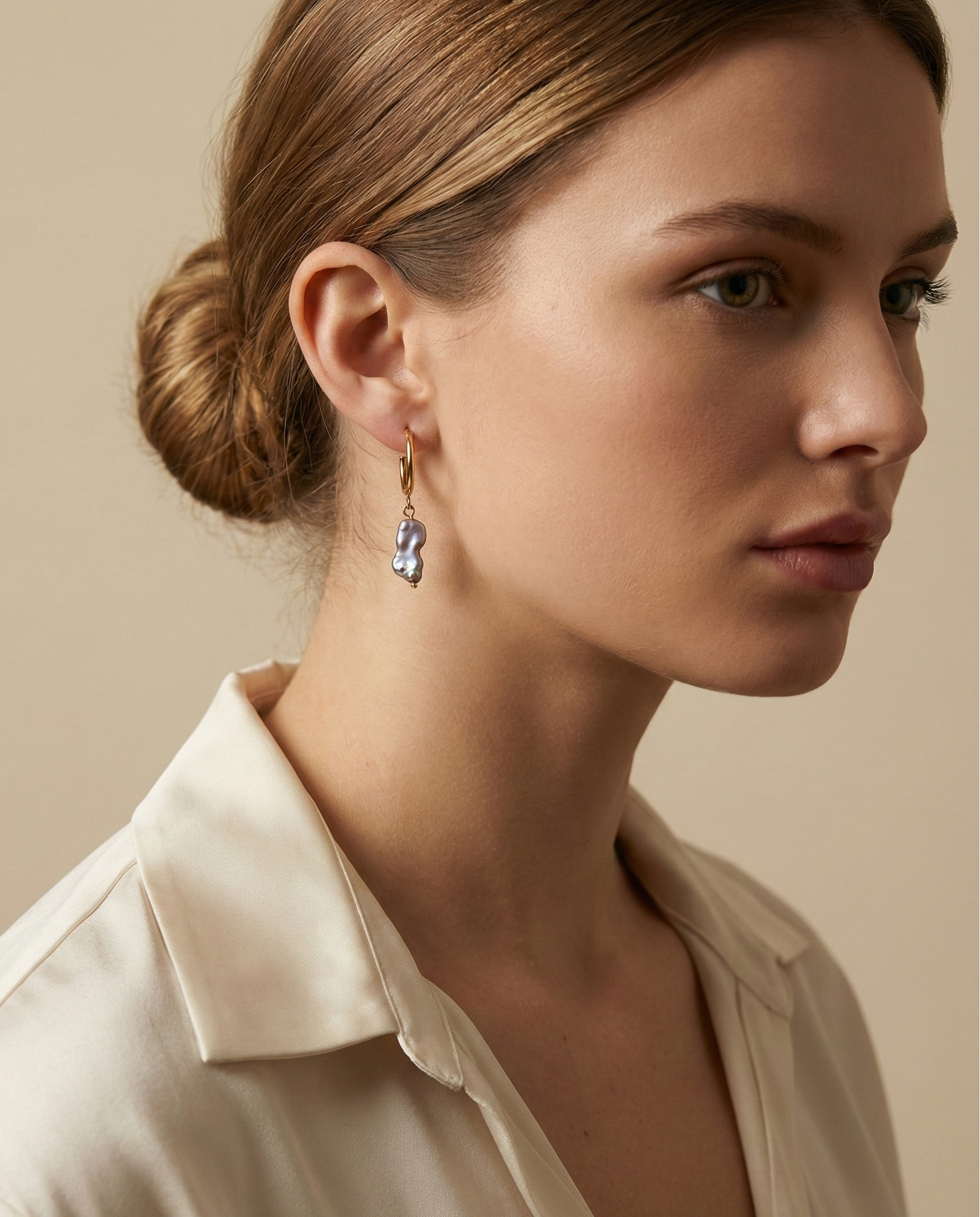 Close-up commercial jewelry photography of a gold huggie hoop earring featuring a lustrous grey keshi pearl drop. The organic, baroque-shaped pearl hangs from a sleek gold ring on a model’s ear, framed by soft blonde-brown hair. The image showcases high-quality nacre iridescence, realistic skin texture, and a warm, matte ivory background, highlighting the minimalist luxury aesthetic