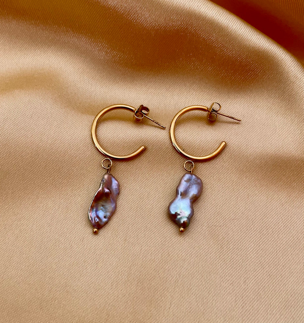 Close-up commercial jewelry photography of a gold huggie hoop earring featuring a lustrous grey keshi pearl drop. The organic, baroque-shaped pearl has high-quality nacre iridescence, realistic skin texture, and a warm, matte ivory background, highlighting the minimalist luxury aesthetic