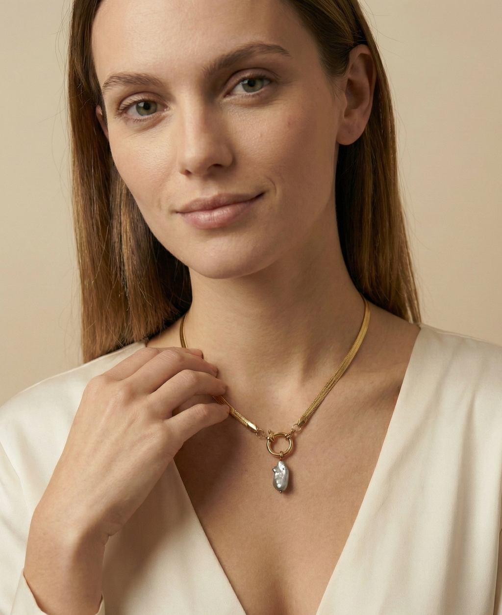 Close-up of a model wearing a flat gold herringbone chain necklace featuring a front toggle clasp and an elongated grey keshi pearl pendant, styled with a deep V-neck ivory silk top