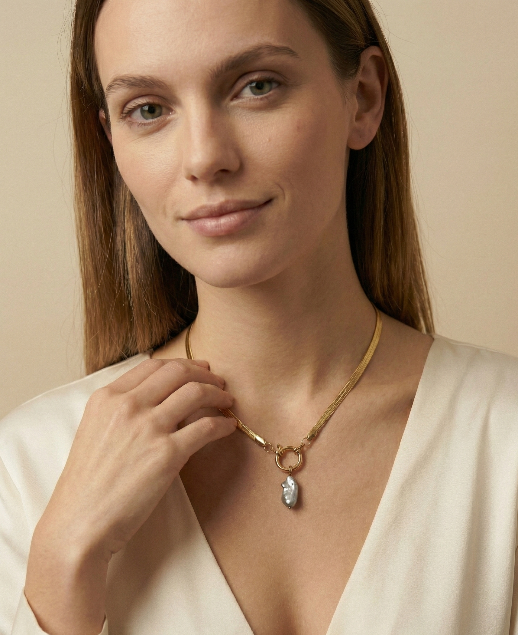 Close-up of a model wearing a flat gold herringbone chain necklace featuring a front toggle clasp and an elongated grey keshi pearl pendant, styled with a deep V-neck ivory silk top