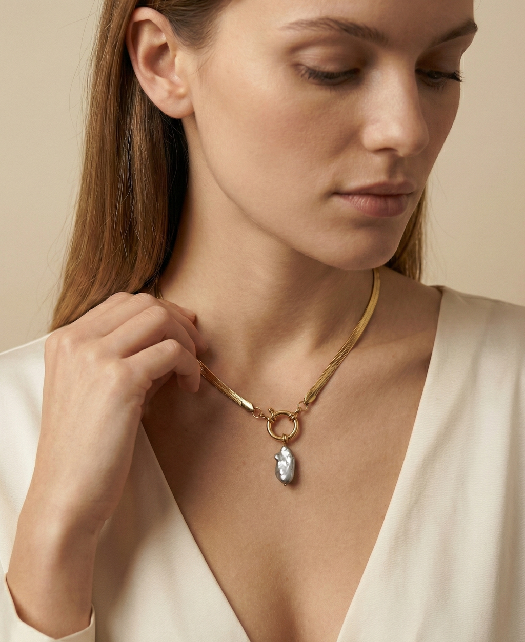 Close-up of a model wearing a flat gold herringbone chain necklace featuring a front toggle clasp and an elongated grey keshi pearl pendant, styled with a deep V-neck ivory silk top