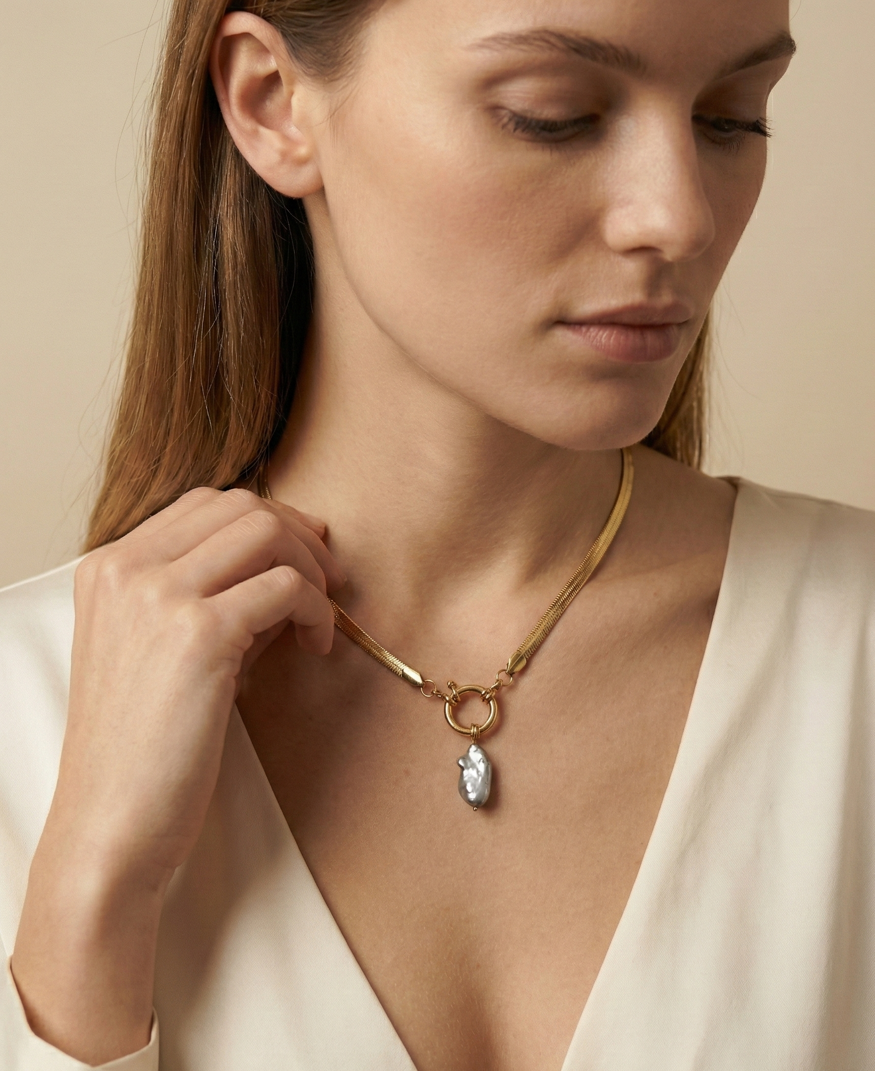 Close-up of a model wearing a flat gold herringbone chain necklace featuring a front toggle clasp and an elongated grey keshi pearl pendant, styled with a deep V-neck ivory silk top