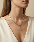 Close-up of a model wearing a flat gold herringbone chain necklace featuring a front toggle clasp and an elongated grey keshi pearl pendant, styled with a deep V-neck ivory silk top