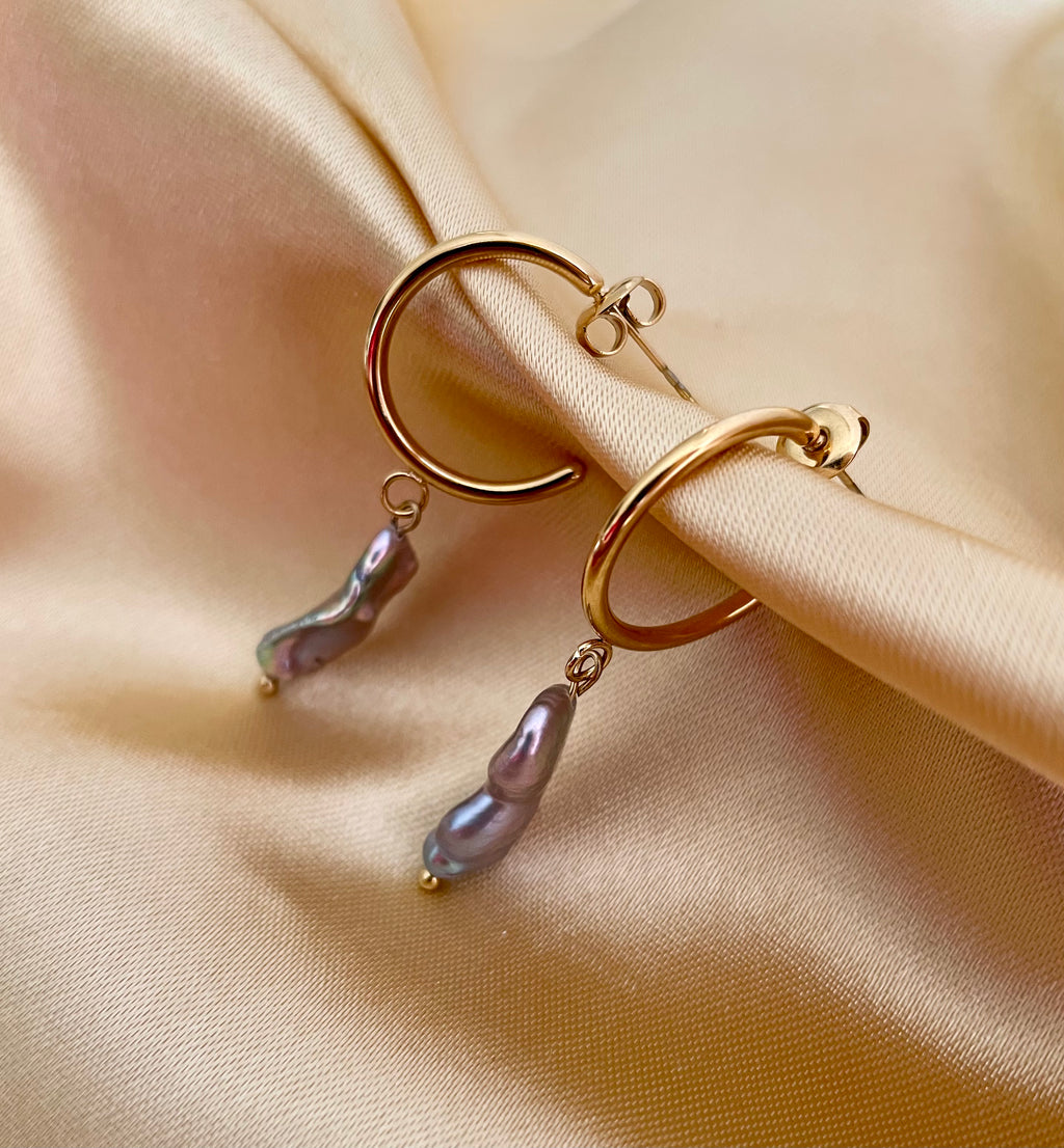 Close-up commercial jewelry photography of a gold huggie hoop earring featuring a lustrous grey keshi pearl drop. The organic, baroque-shaped pearl has high-quality nacre iridescence, realistic skin texture, and a warm, matte ivory background, highlighting the minimalist luxury aesthetic
