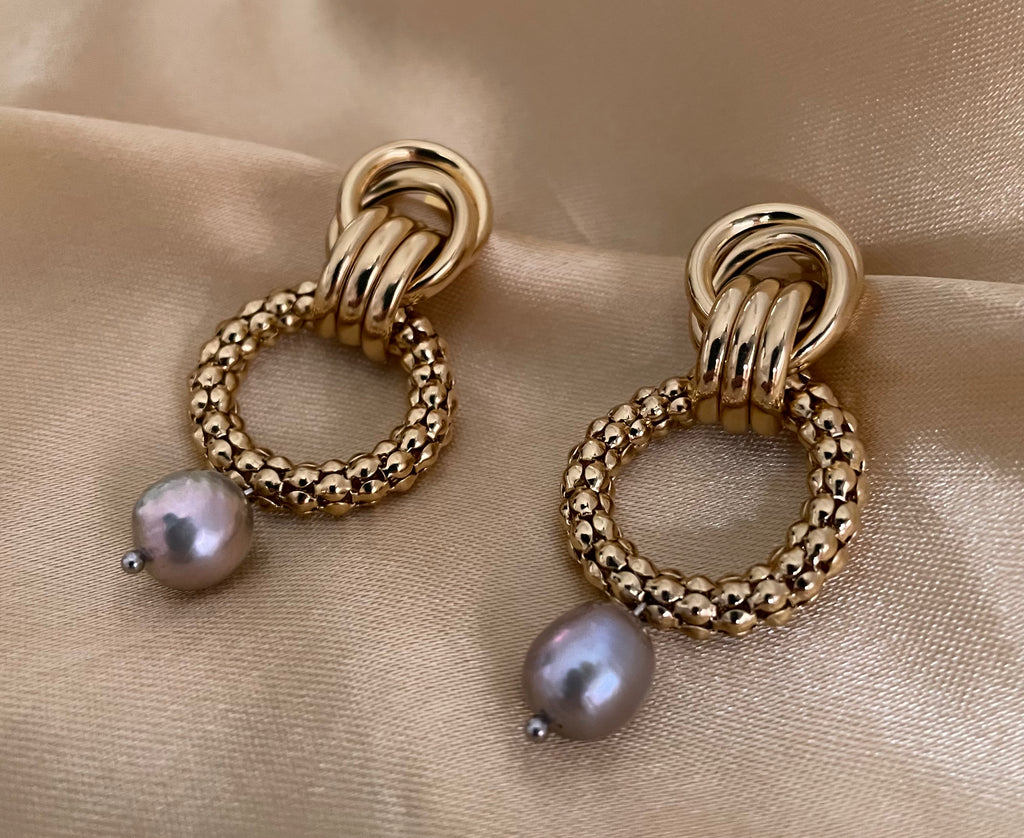 Close-up detail of textured gold interlocking hoop earrings featuring a lustrous metallic grey pearl drop. The design includes a beaded gold circle and polished gold links