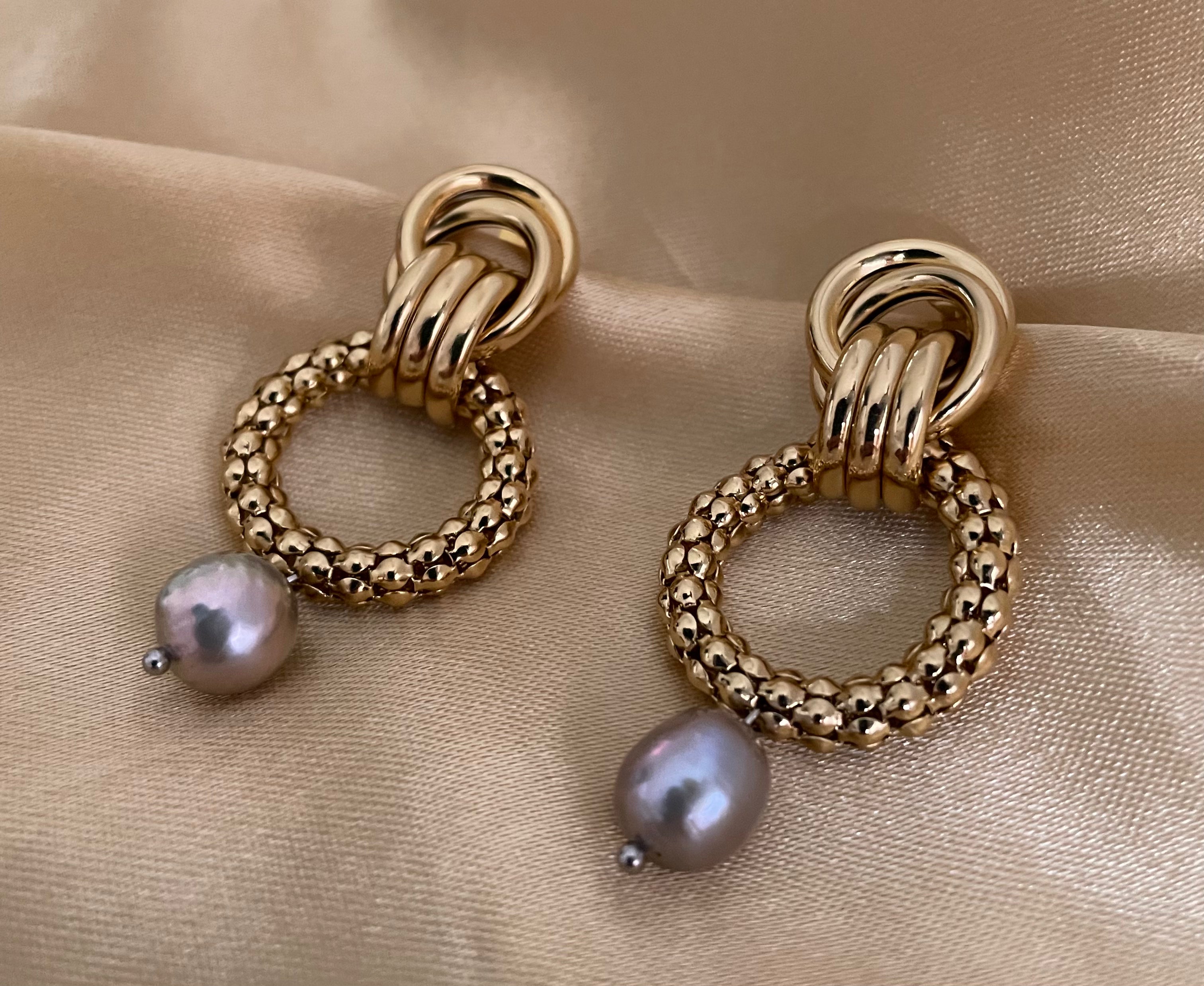 Close-up detail of textured gold interlocking hoop earrings featuring a lustrous metallic grey pearl drop. The design includes a beaded gold circle and polished gold links