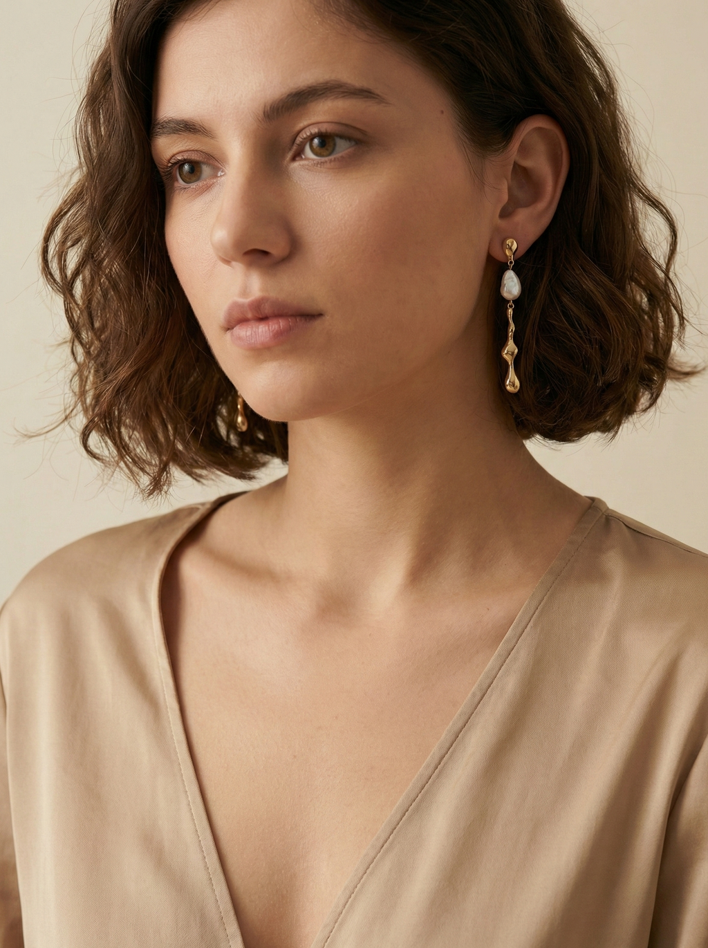 Woman wearing white kasha pearl and gold earrings with a beige top against a neutral background
