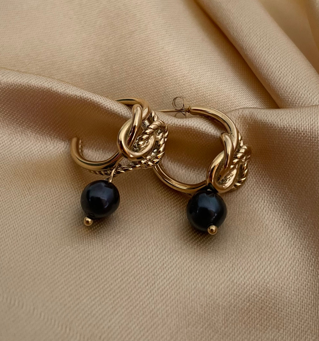 Luxury gold knot hoop earrings with round black pearl drop. High-fashion jewelry photography showing elegant styling with an off-shoulder black top and classic hairstyle. Perfect for evening wear and sophisticated looks