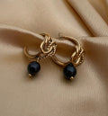 Luxury gold knot hoop earrings with round black pearl drop. High-fashion jewelry photography showing elegant styling with an off-shoulder black top and classic hairstyle. Perfect for evening wear and sophisticated looks