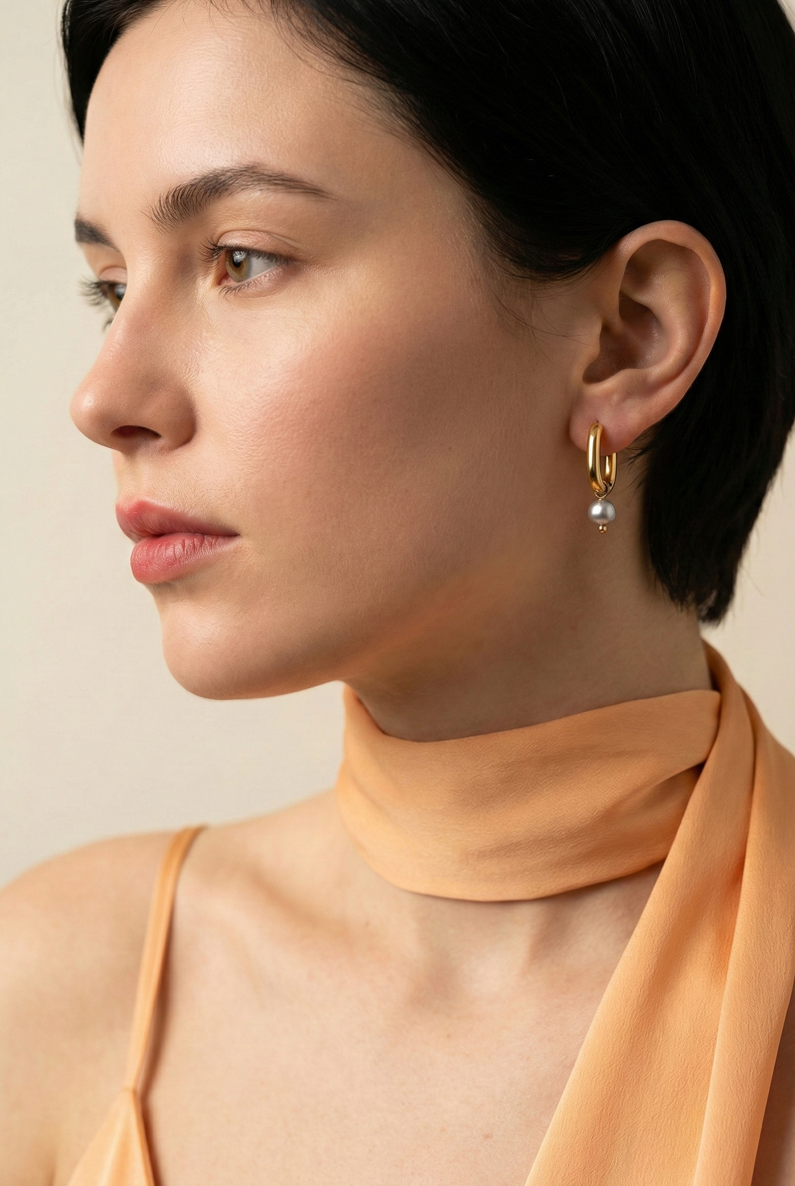 Editorial jewelry portrait showing a side profile of a model wearing luxury gold pearl earrings styled with an orange silk scarf.