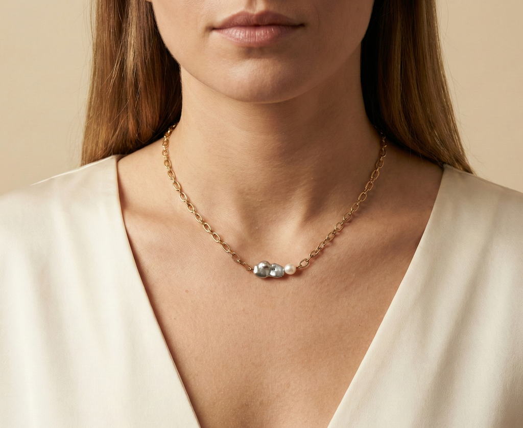 Close-up editorial portrait of a woman wearing a gold oval-link chain necklace featuring a grey baroque keshi pearl and a small white pearl, styled with a deep V-neck ivory silk top