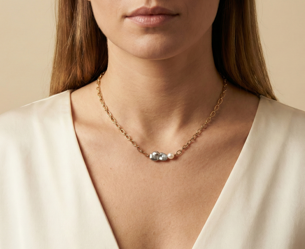Close-up editorial portrait of a woman wearing a gold oval-link chain necklace featuring a grey baroque keshi pearl and a small white pearl, styled with a deep V-neck ivory silk top