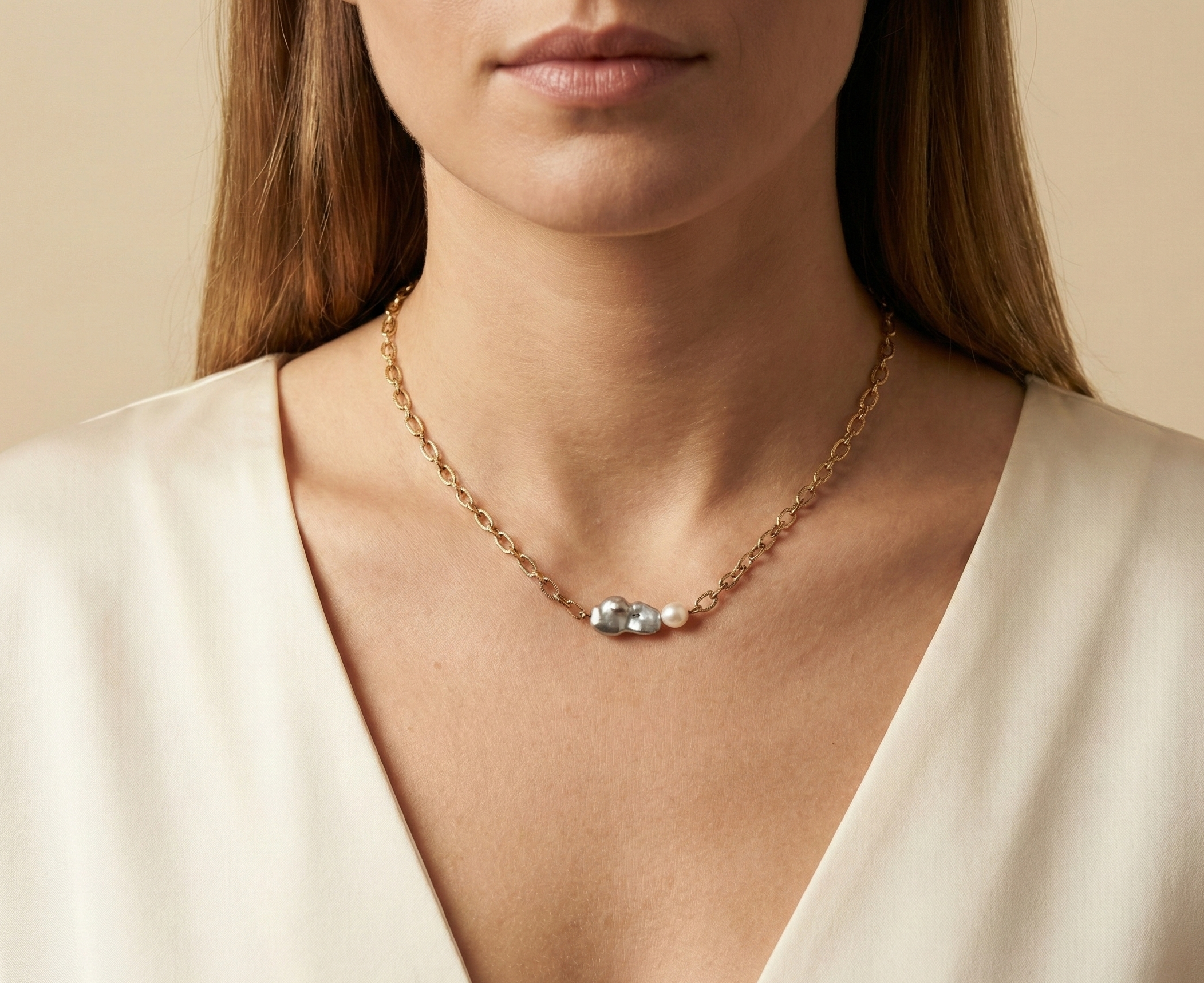 Close-up editorial portrait of a woman wearing a gold oval-link chain necklace featuring a grey baroque keshi pearl and a small white pearl, styled with a deep V-neck ivory silk top