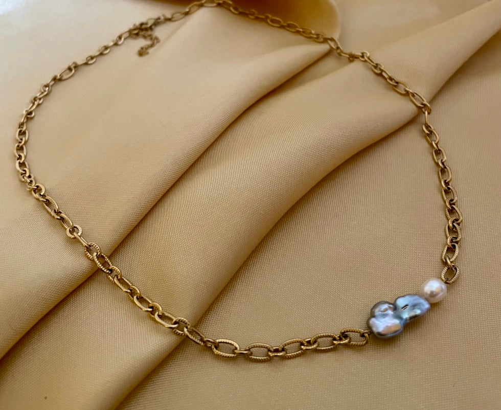 Close-up editorial portrait of a woman wearing a gold oval-link chain necklace featuring a grey baroque keshi pearl and a small white pearl, styled with a deep V-neck ivory silk top