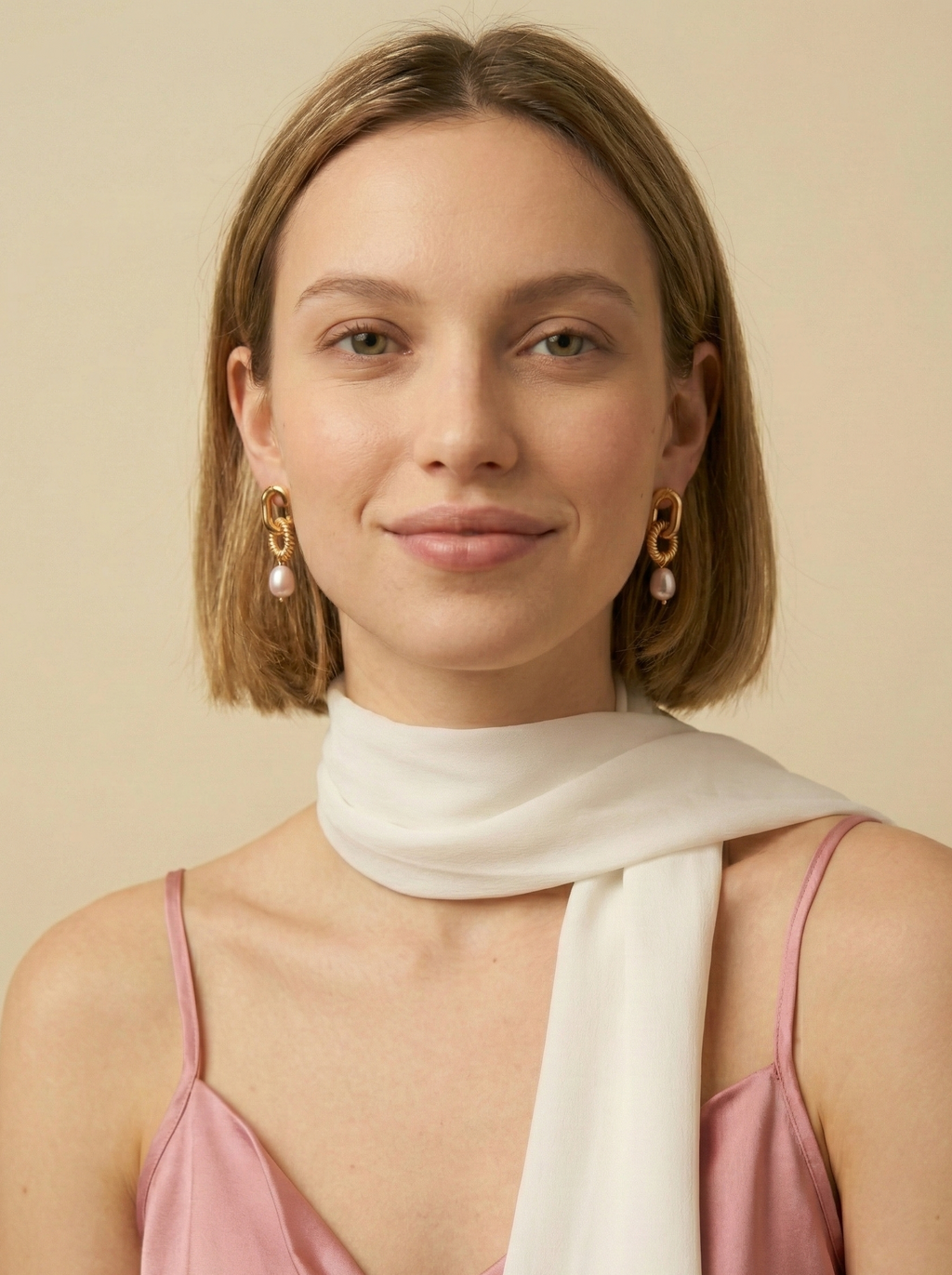 A female model wearing chunky gold interlocking link earrings with pearl drops. The vintage-inspired jewelry creates an elegant statement against a soft ivory background, styled with a pink silk camisole and white scarf