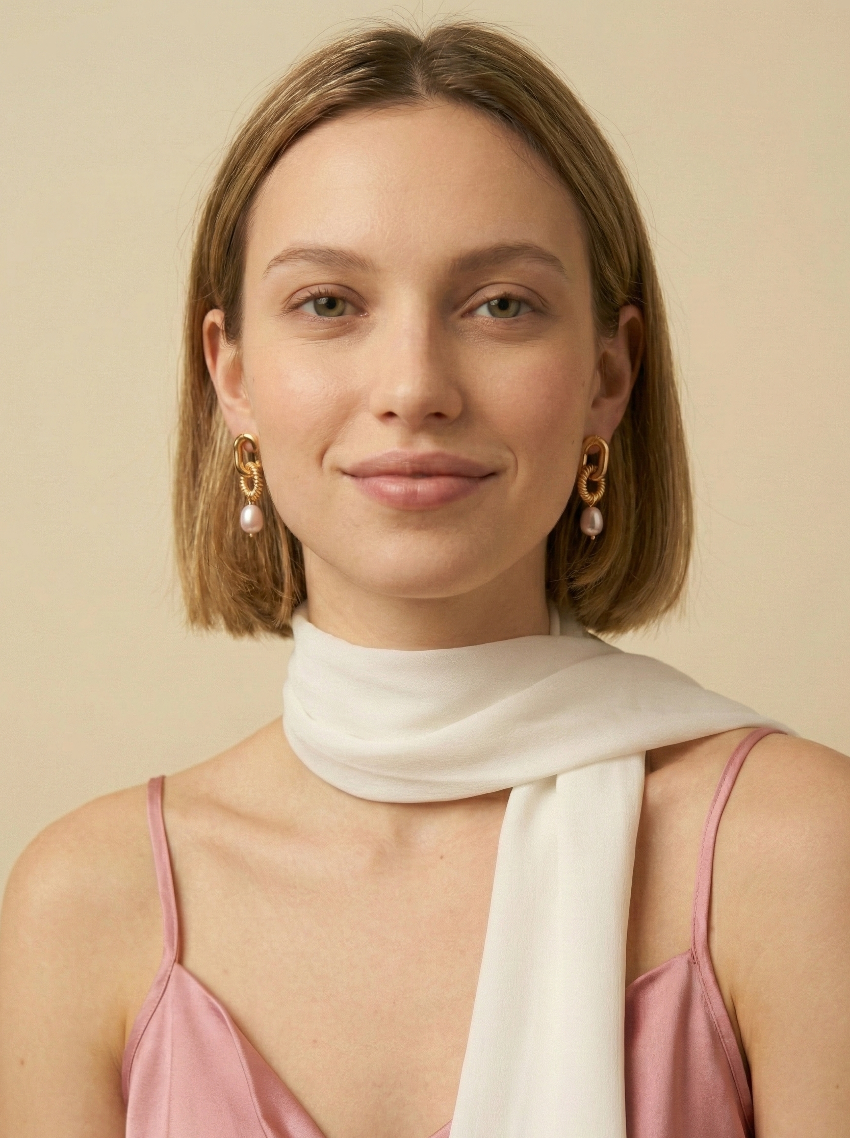 A female model wearing chunky gold interlocking link earrings with pearl drops. The vintage-inspired jewelry creates an elegant statement against a soft ivory background, styled with a pink silk camisole and white scarf