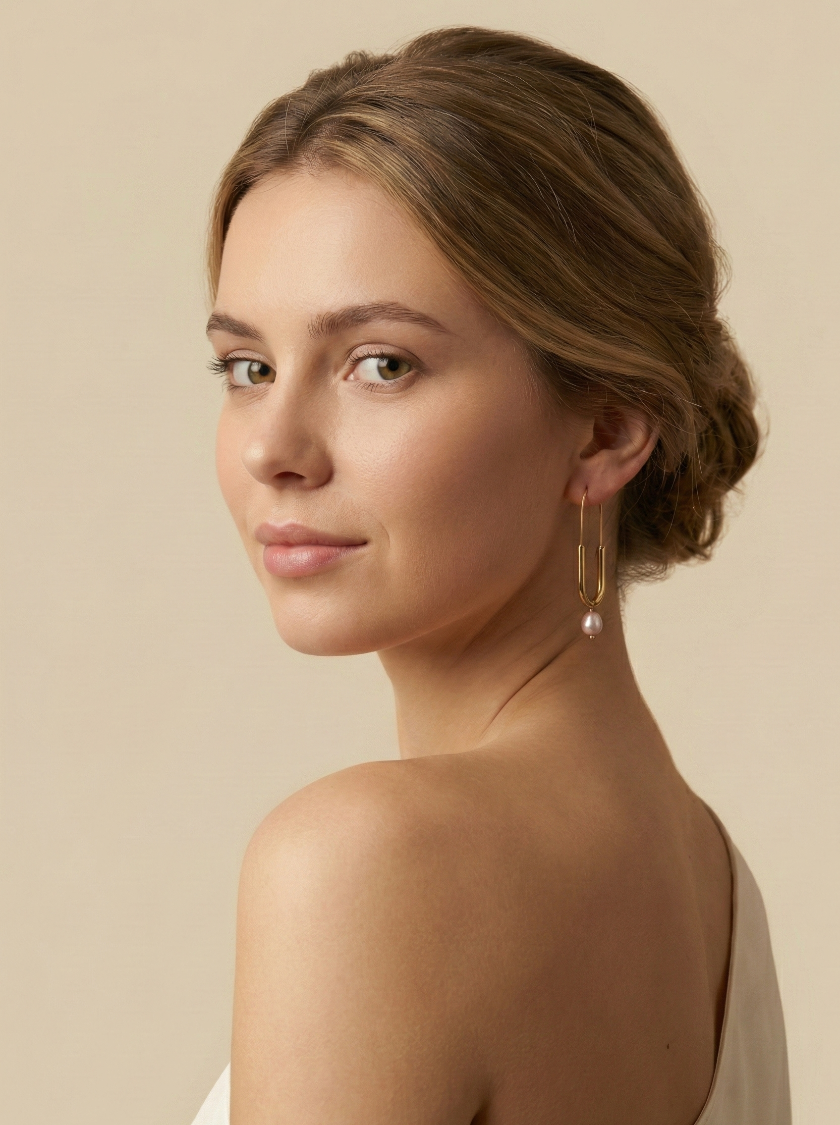 Close-up of a model wearing modern gold safety pin drop earrings featuring a lustrous pink oval pearl charm. Minimalist gold wire earrings with a U-shape design and natural pearl detail