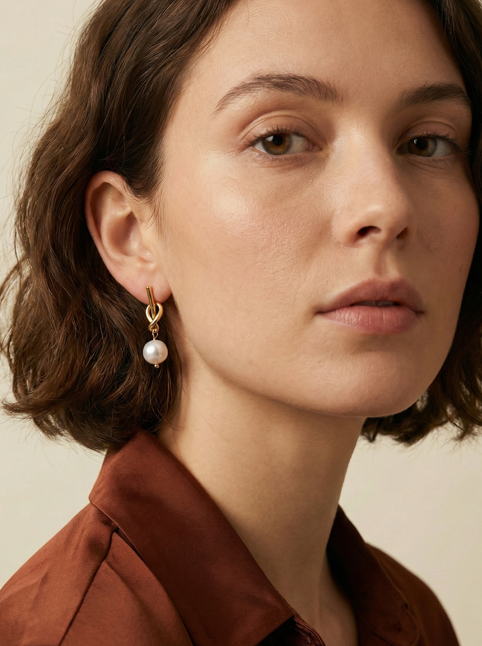 Close-up of a model wearing gold drop earrings featuring a unique knot design and a dangling white round pearl. The earrings are styled with a rust-colored silk shirt