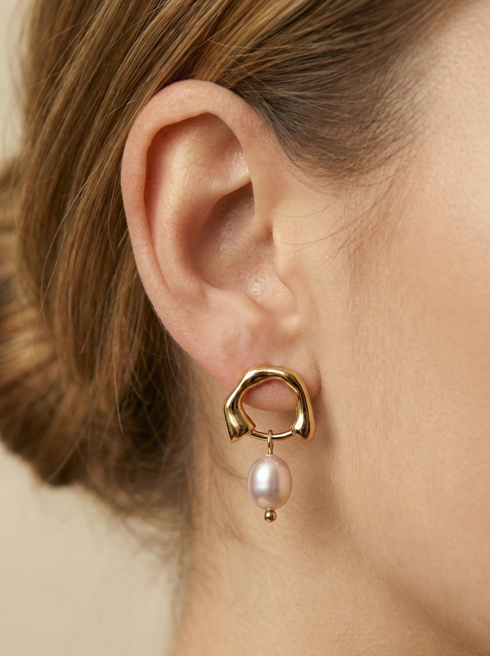 Close-up detail of organic gold huggie hoop earrings featuring a high-luster pink oval freshwater pearl drop, worn on a model's ear against a matte ivory background