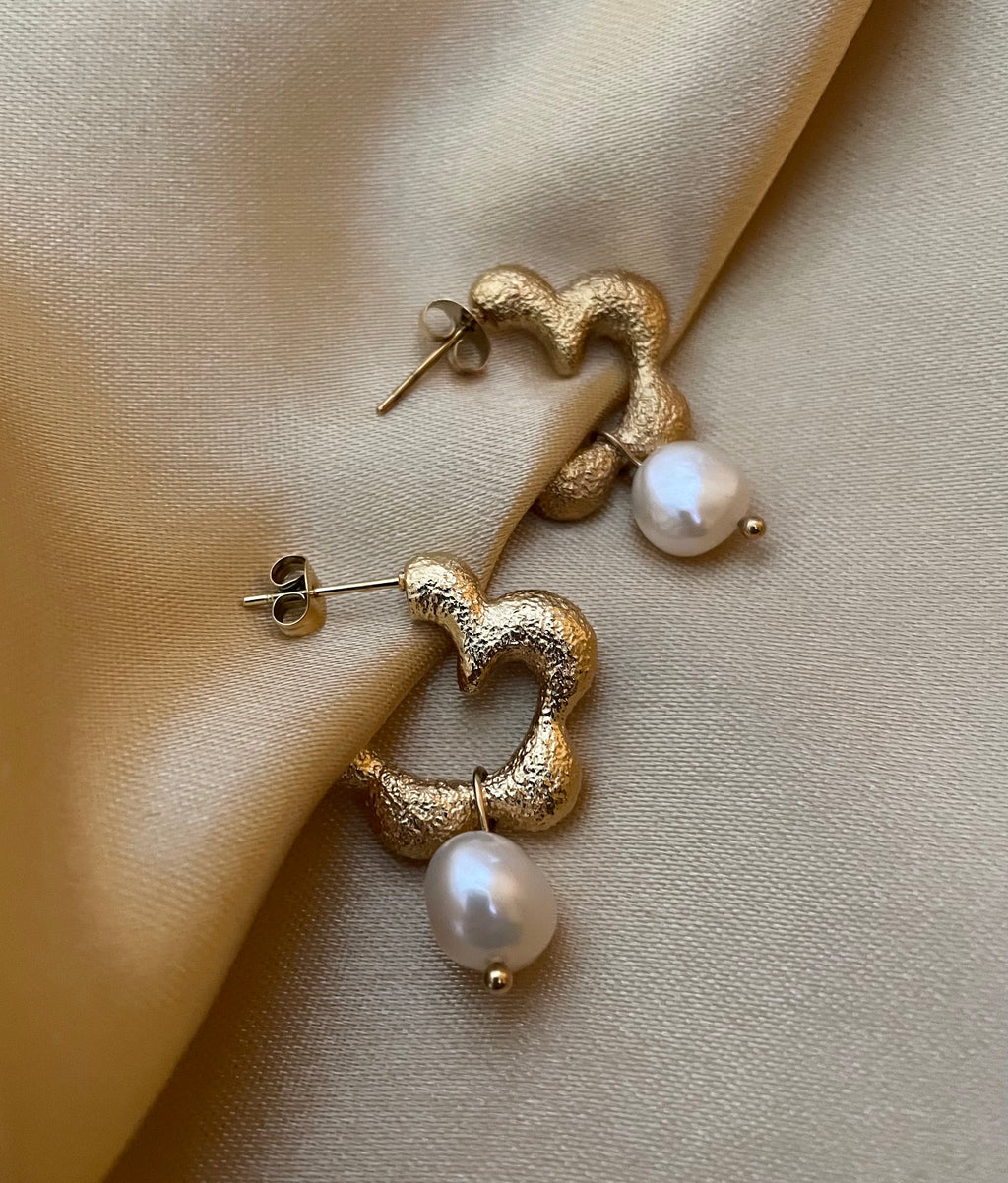 Close-up of textured gold huggie hoop earrings featuring a lustrous white pearl drop. The earrings have an organic, hammered gold finish