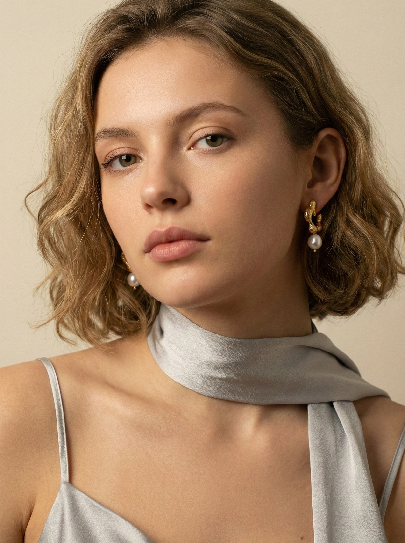 Model wearing small textured gold hoop earrings with a single round white pearl drop and a grey silk neck scar