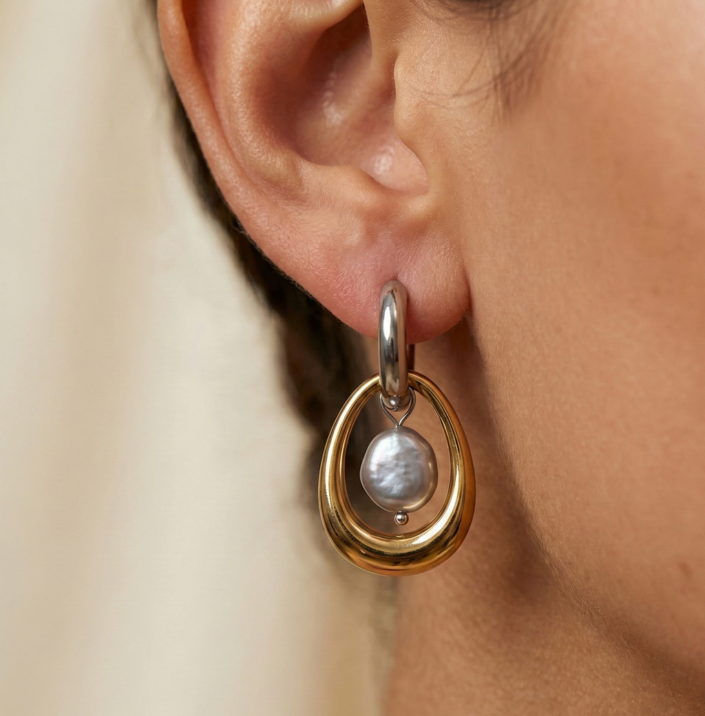 Gold and silver earring with a baroque round grey pearl drop worn by a person.