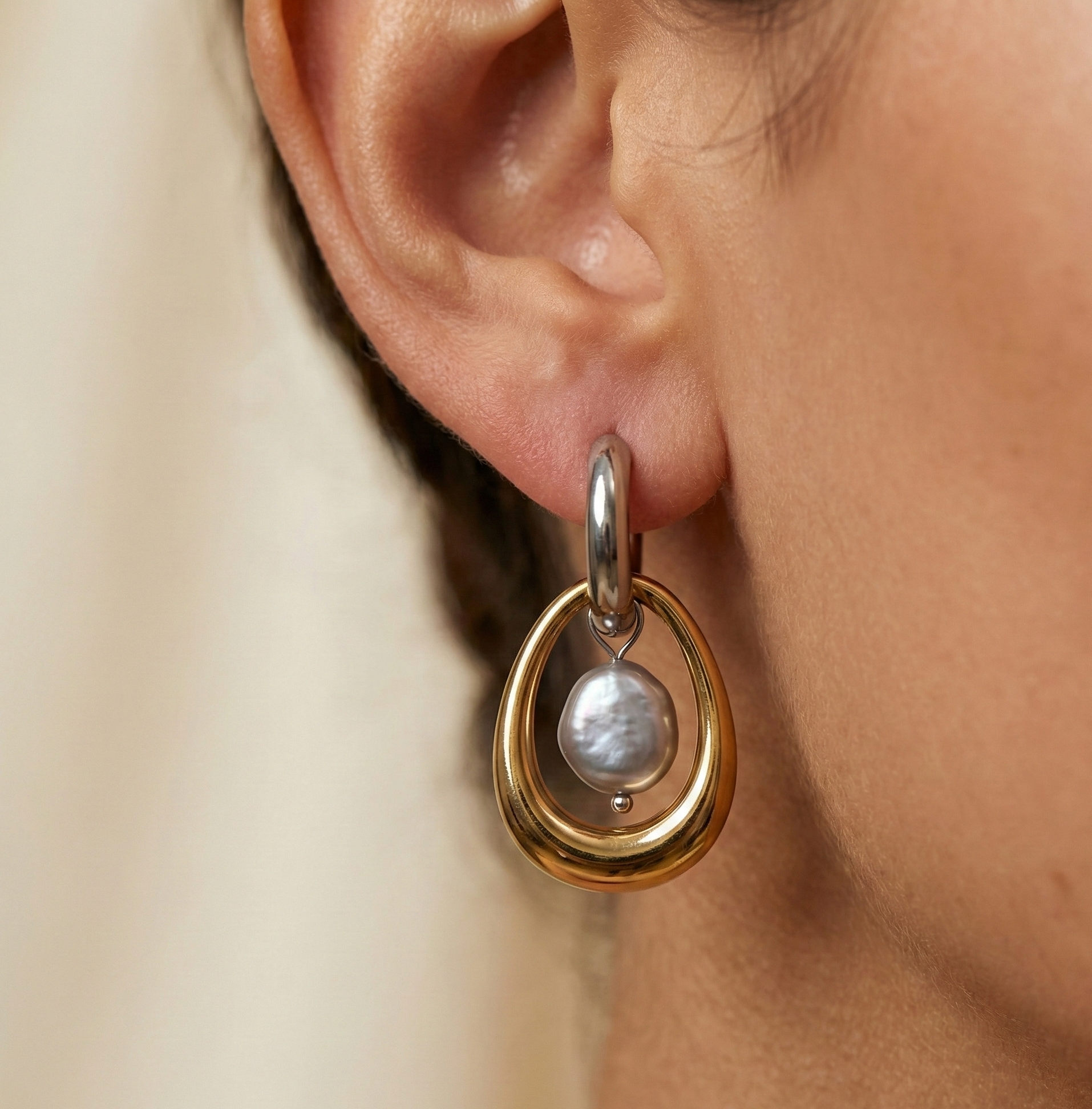 Gold and silver earring with a baroque round grey pearl drop worn by a person.