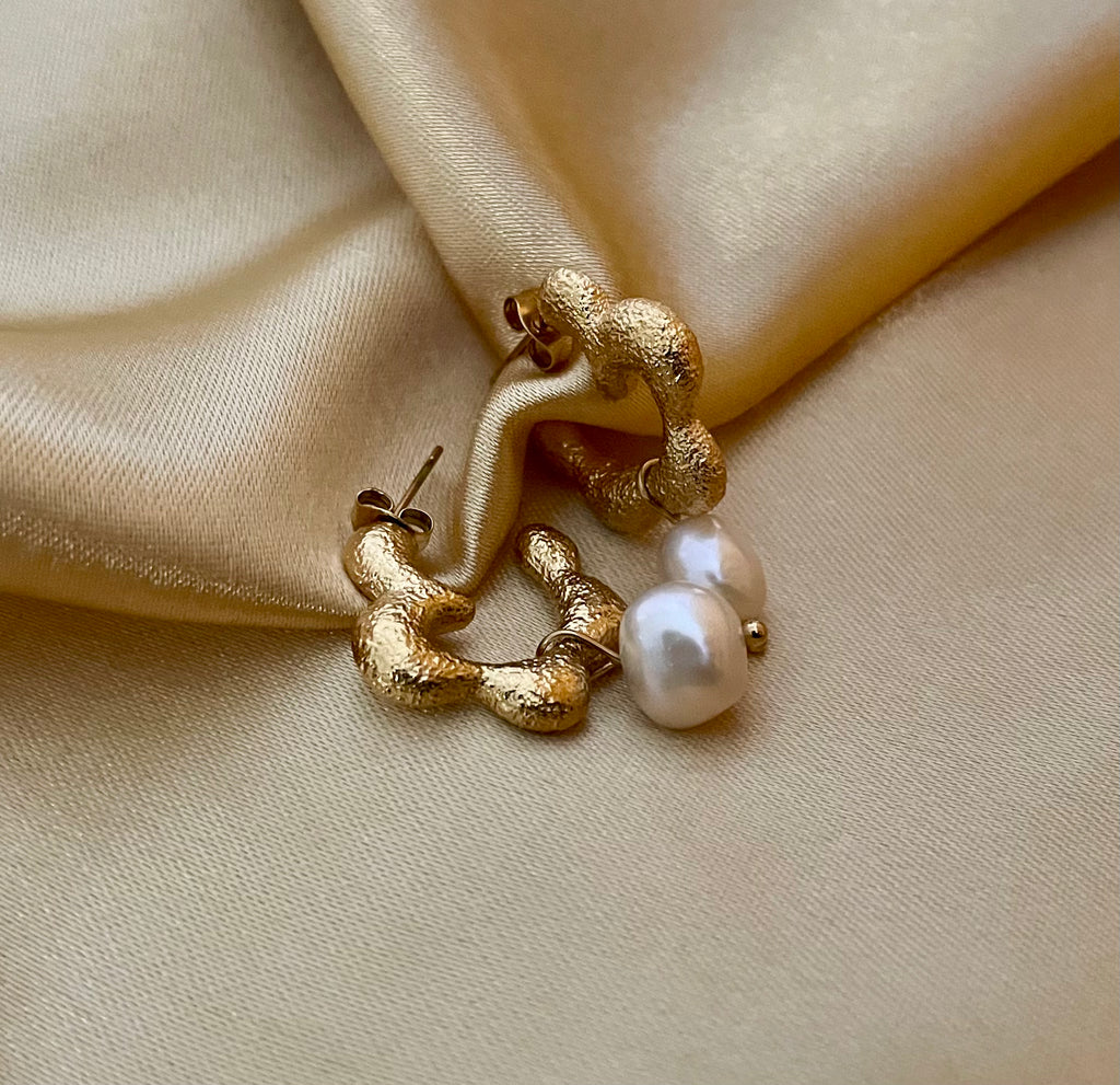 Close-up of textured gold huggie hoop earrings featuring a lustrous white pearl drop. The earrings have an organic, hammered gold finish