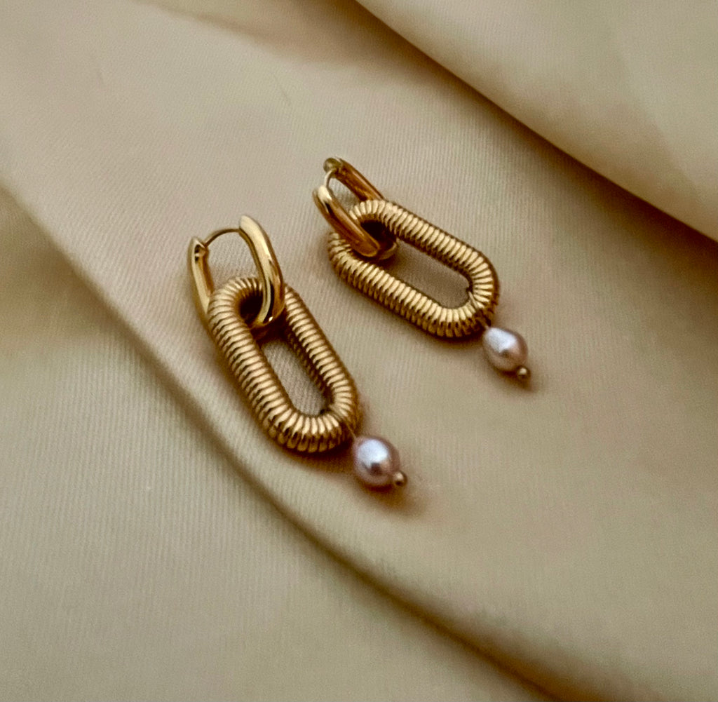 Close-up of textured gold oval link earrings featuring a dainty pink pearl drop, worn on a model's ear. High-resolution commercial jewelry photography showcasing ribbed gold detailing and lustrous pearl against a soft matte ivory background.
