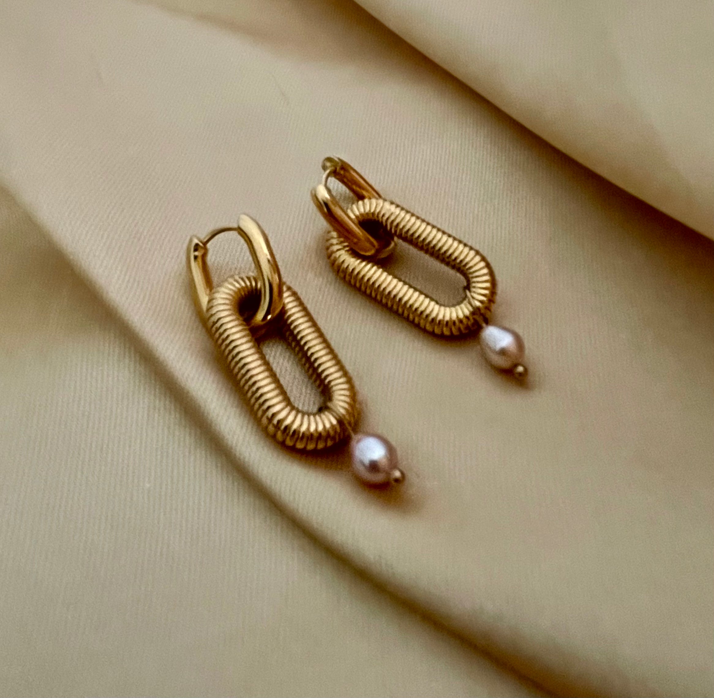 Close-up of textured gold oval link earrings featuring a dainty pink pearl drop, worn on a model's ear. High-resolution commercial jewelry photography showcasing ribbed gold detailing and lustrous pearl against a soft matte ivory background.