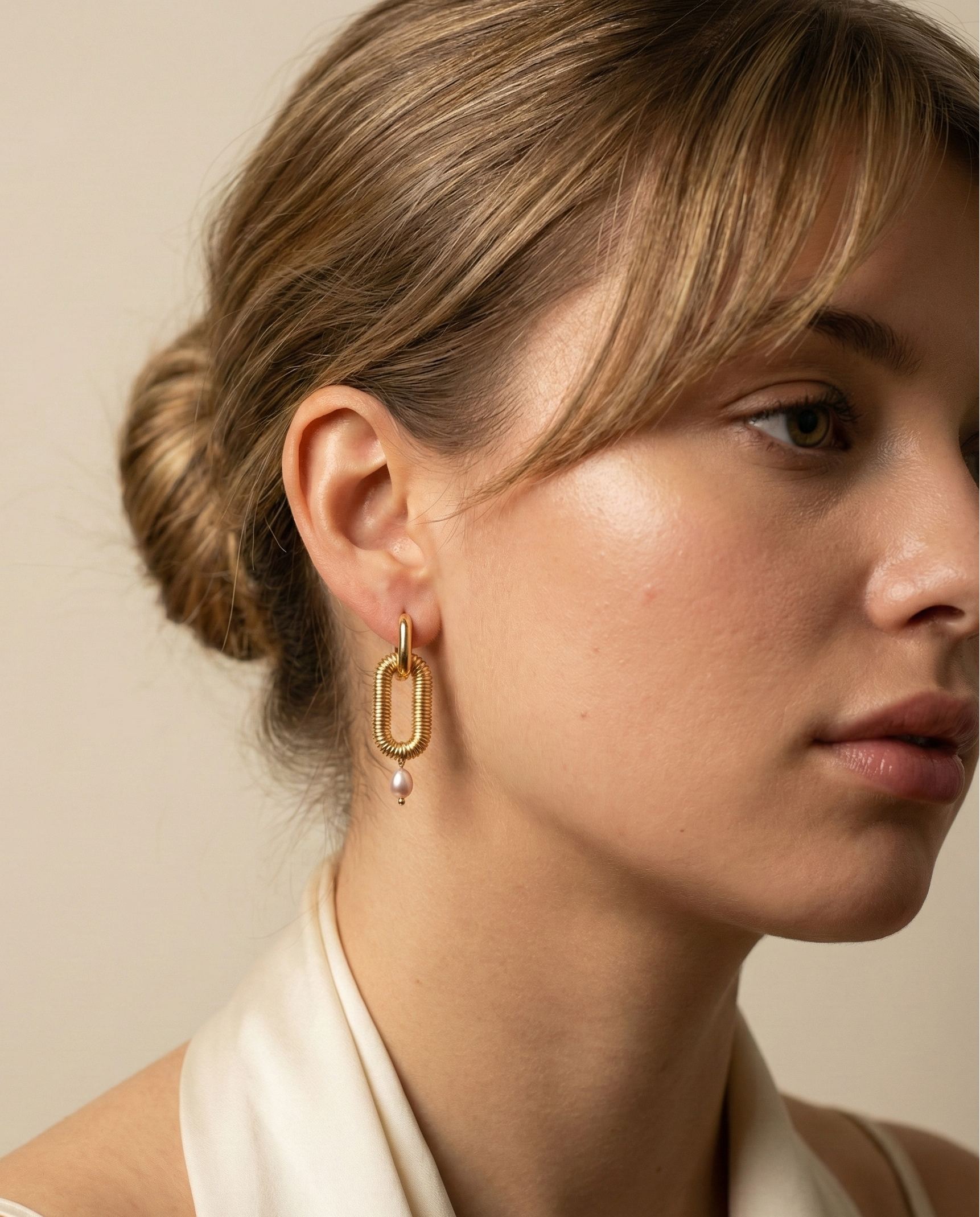 Close-up of textured gold oval link earrings featuring a dainty pink pearl drop, worn on a model's ear. High-resolution commercial jewelry photography showcasing ribbed gold detailing and lustrous pearl against a soft matte ivory background.