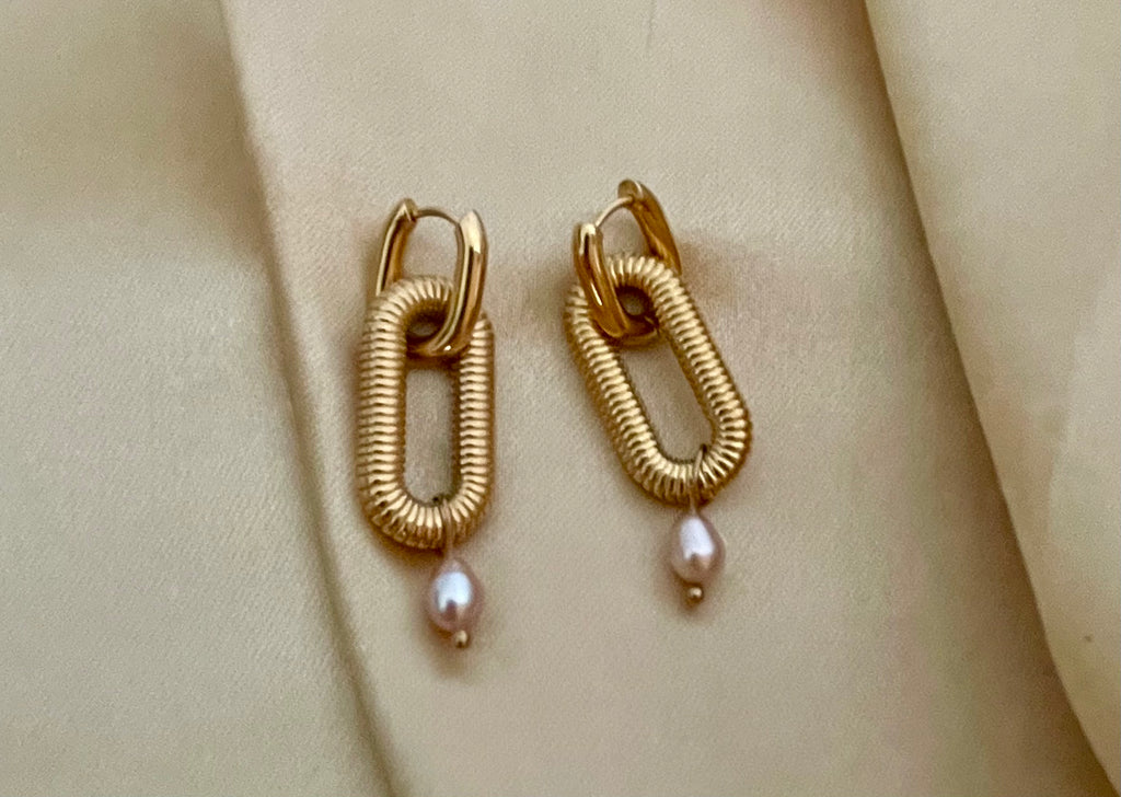 Close-up of textured gold oval link earrings featuring a dainty pink pearl drop, worn on a model's ear. High-resolution commercial jewelry photography showcasing ribbed gold detailing and lustrous pearl against a soft matte ivory background.