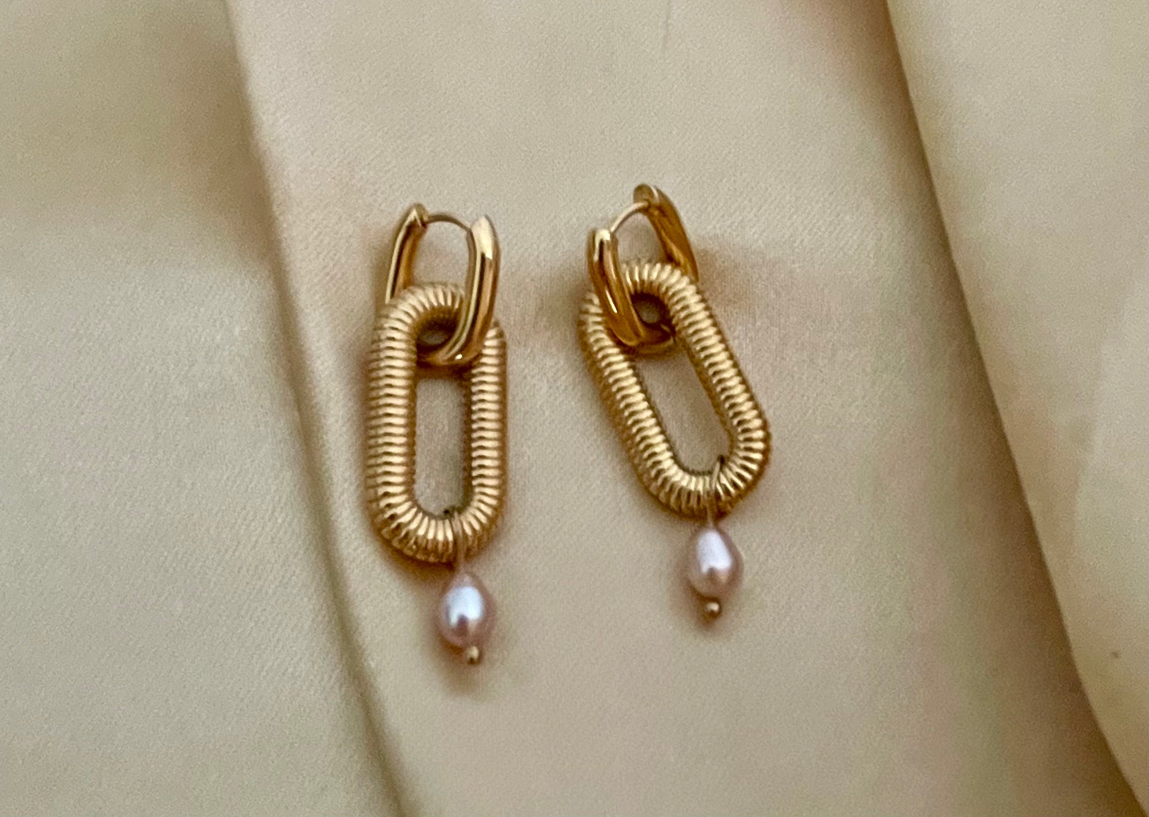 Close-up of textured gold oval link earrings featuring a dainty pink pearl drop, worn on a model's ear. High-resolution commercial jewelry photography showcasing ribbed gold detailing and lustrous pearl against a soft matte ivory background.