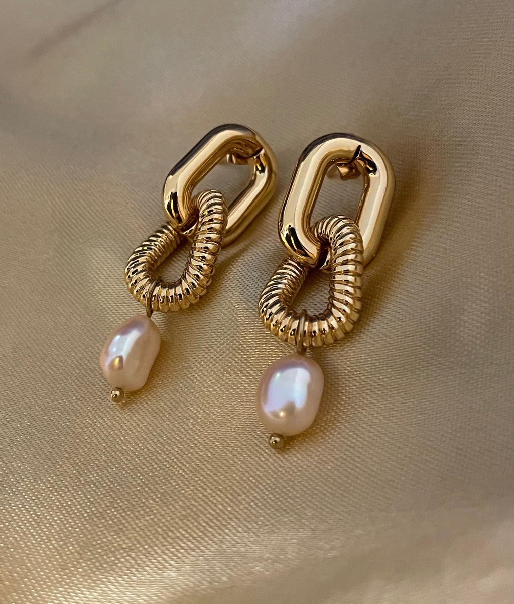 Close-up of sculptural gold link earrings featuring a twisted rope texture and hanging freshwater baroque pearl drops. The earrings are displayed on a champagne silk fabric to highlight the pearl luster and high-shine gold finish