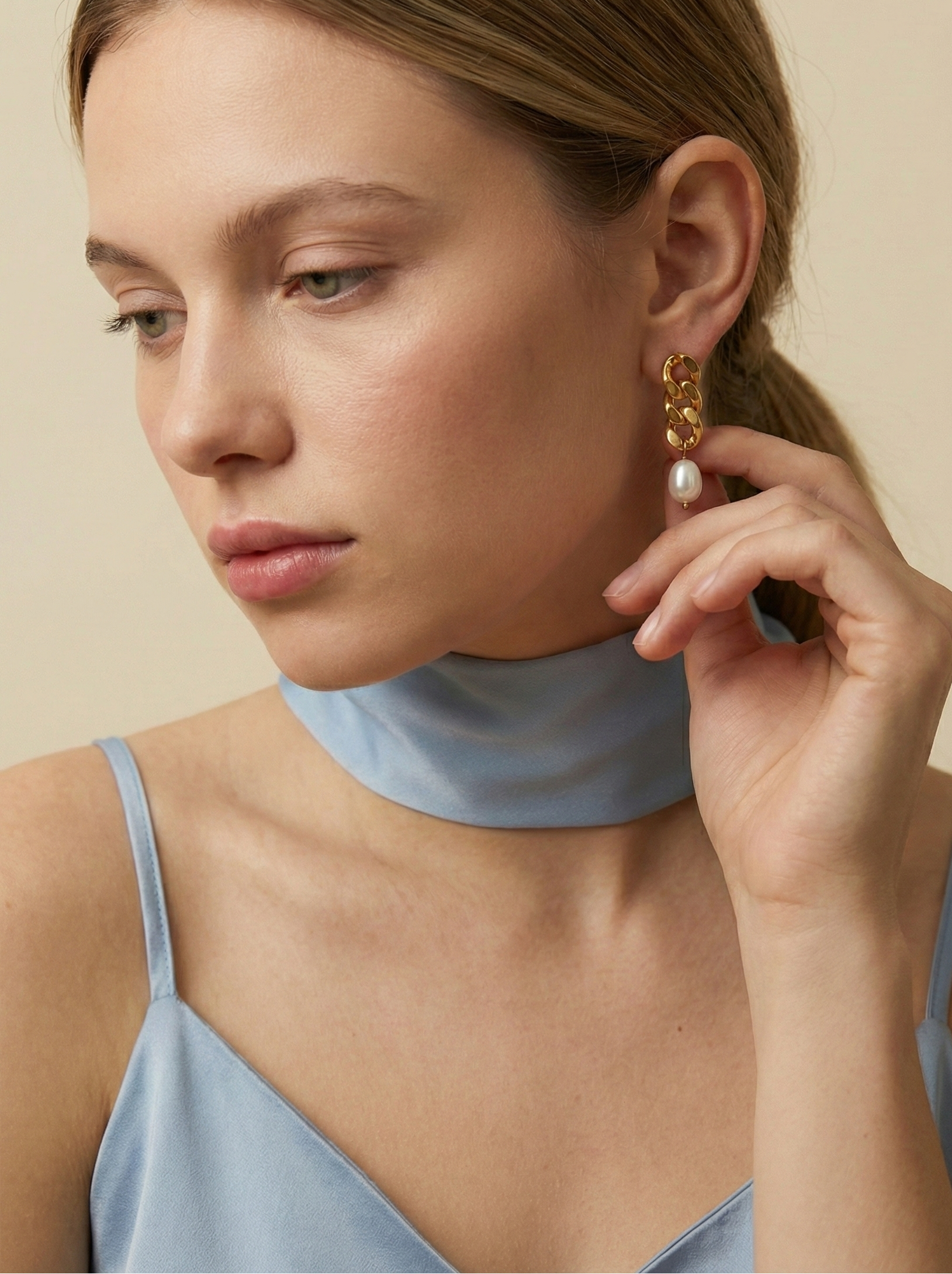 Editorial beauty portrait of a model wearing chunky gold link pearl earrings paired with a light blue silk scarf. The image highlights luxury jewelry trends and elegant styling