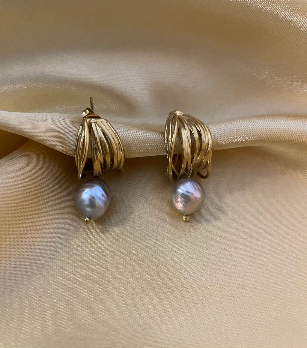 Luxury fashion jewelry photography of gold twist hoop earrings with dangling grey pearls. High-resolution close-up on the ear showing realistic skin texture, intricate gold detailing, and a sophisticated silver outfit.